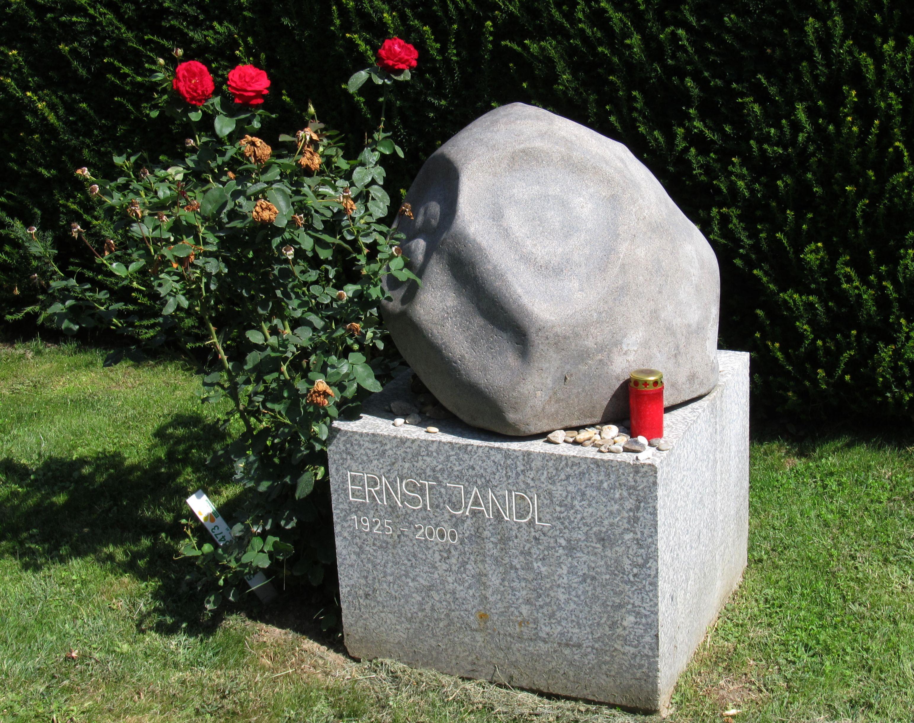 Friedhofsszene. Auf einem quadratischen Marmorsockel, auf dem steht: ERNST JANDL 1925 - 2000,  liegt ein großer, weißer Stein, mit rundgeschliffenen Kanten. Daneben auf dem grünem Rasen ein Rosenbusch mit ein paar roten Blüten. Auf dem Marmorblock liegen rechts und links viele kleine Kieselsteine, rechts an der Ecke steht eine rote Kerze. Im Hintergrund verläuft eine grüne Hecke.