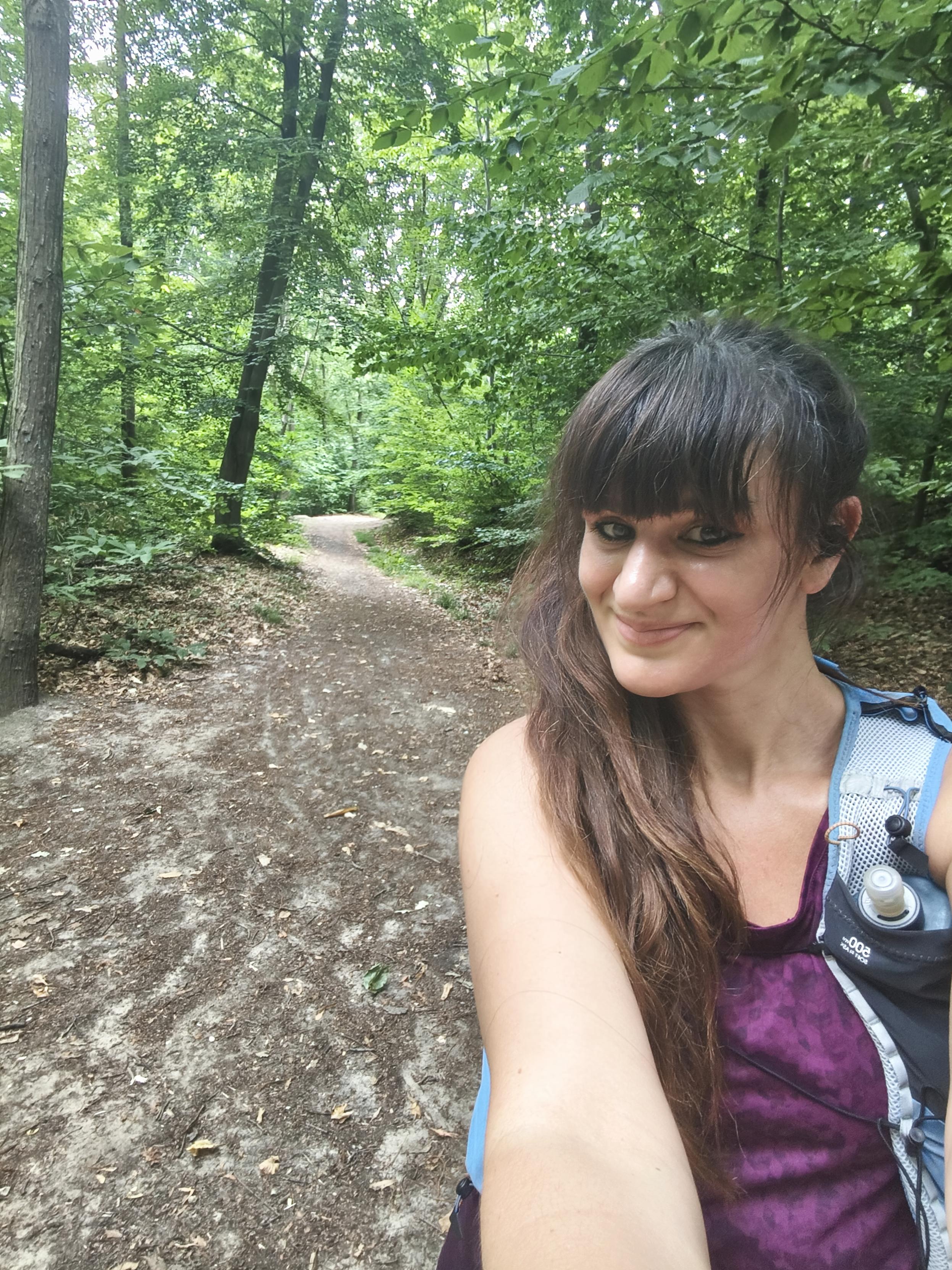 Selfie d'une coureuse souriante sur un large chemin forestier. Elle porte un débardeur violet et un gilet d'hydratation, avec une gourde visible sur la poitrine. Derrière elle, le sentier disparaît sous les grands arbres aux feuillages denses, offrant une impression de fraîcheur malgré la chaleur de l'été. 🌿🏃‍♀️