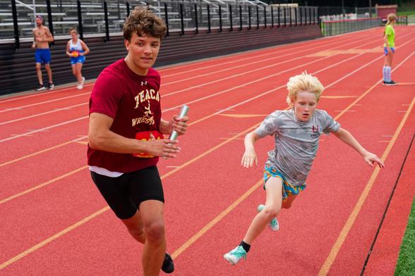 Young man with maroon colored shirt is holding an aluminum-colored cylinder that was just handed to him by a small boy running to the right