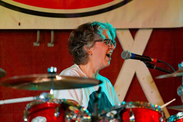 Female drummer in white shirt with short hair sings into microphone.