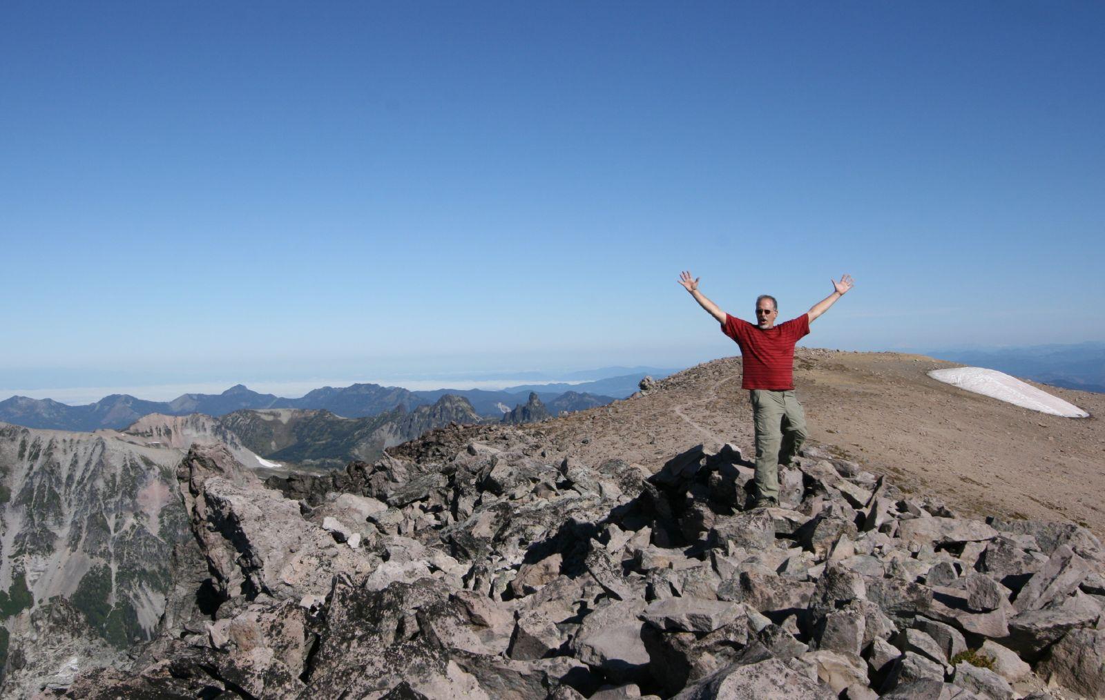 Hiker in red shirt and green pants with arms spread up at an angle howling something like "Wow!"  from a pile of rocks at the top of a high hill, with the trail leading to it visible in the background. The sky is pale blue and you can see more mountains in the distance.