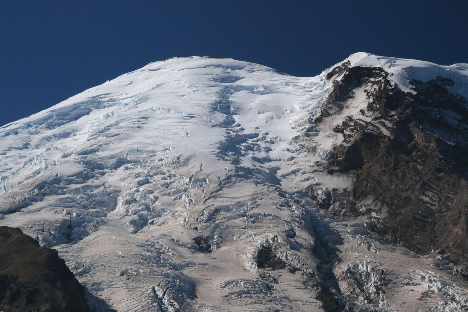 As seen from the top of the associated Third Burroughs Mountain, the white ice of the Emmons glacier descends from the summit of Tahoma, or Mount Rainier. You can see many cracks and crevasses in the huge glacier, with a huge rock ridge on the right. Sky above the rounded dome of the mountain is pretty blue.