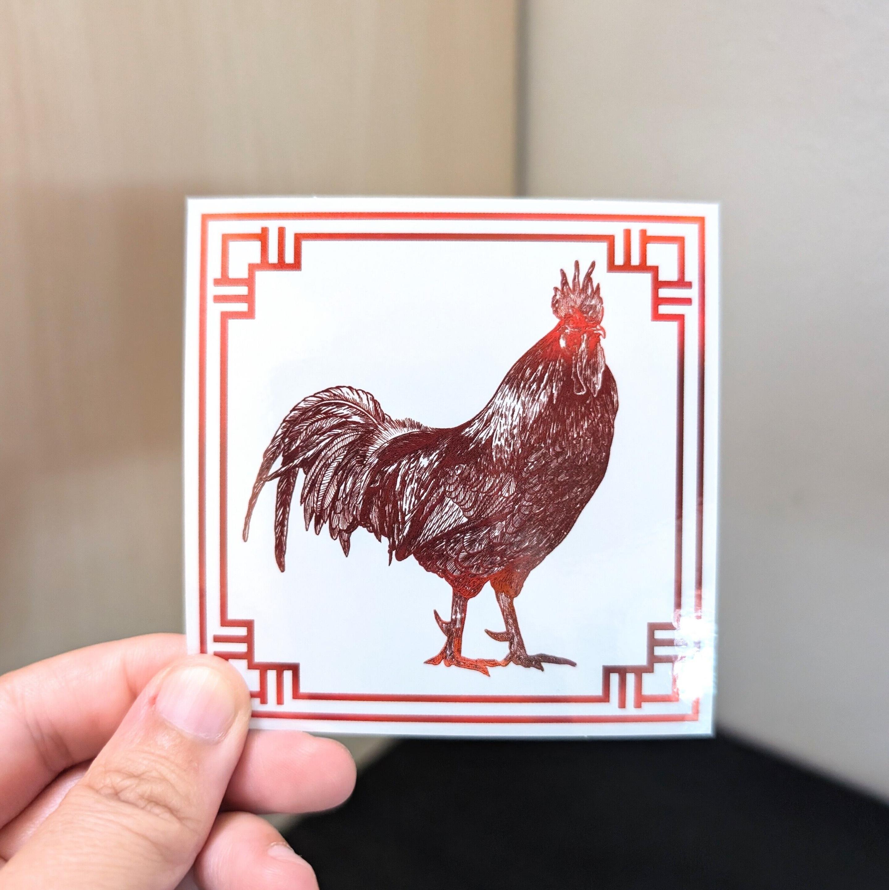 Photo of a left hand holding a square sticker. The sticker is a holographic drawing of the Chinese Zodiac animal: the Rooster.