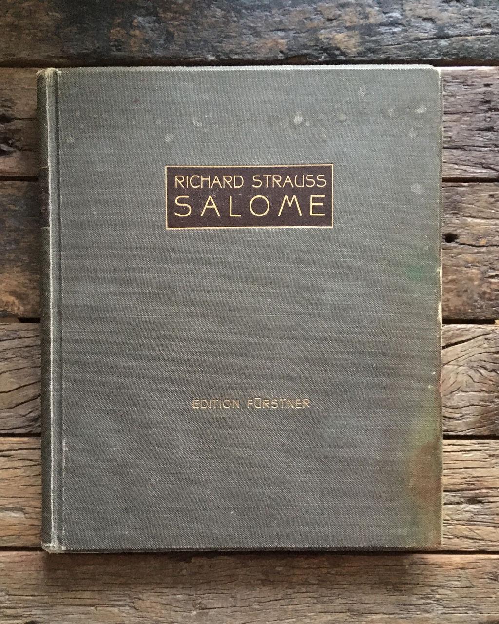 Old and worn piano reduction of the opera Salome by Richard Strauss bound in gray-green linen lies on a rustic wooden table. The title reads
RICHARD STRAUSS
SALOME
in white on black, the name of the imprint
EDITION FÜRSTNER
is debossed in gold.