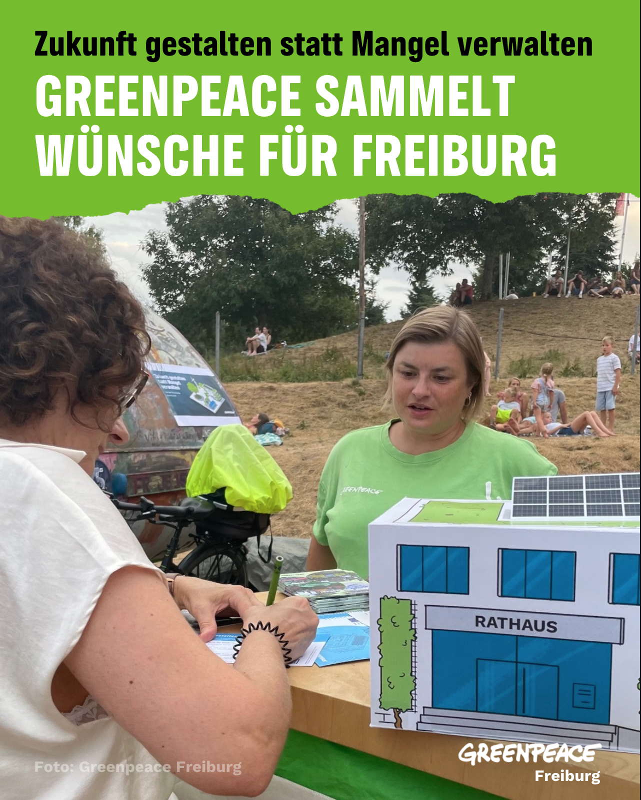 Greenpeace Infostand auf Musikfestival in Freiburg. eine ehrenamtliche ist zu sehen, sowie eine Frau, die etwas ausfüllt. Auf dem Tisch steht ein kleines Rathaus aus Papier. Bürger:innen konnten auf symbolische 100-Millionen-Euro-Scheine schreiben, wofür Freiburg dringend mehr Geld benötigt.