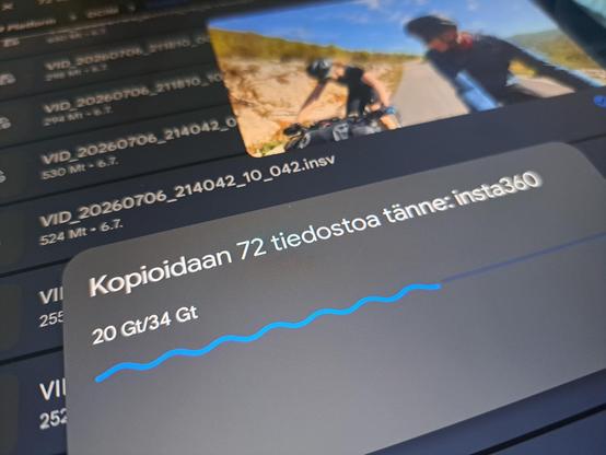 Kuva Android laitteen ruudulta, jossa on kopiointidialogi "Kopioidaan 72 tiedostoa" yhteensä kymmeniä gigatavuja. Dialogin vieressä pienessä ikkunassa pyörii pyöräilyvideo.