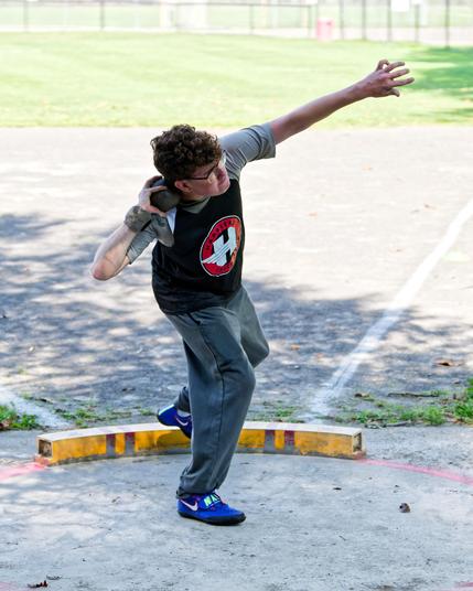 Boy holds a shot put against his neck with one foot on the grond with his arm stretched out ready to toss it.