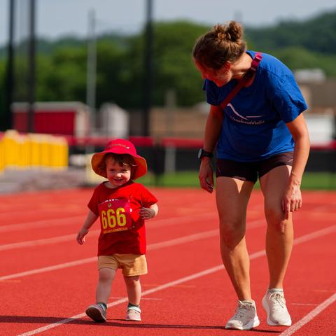 A toddler with a red hat and red shirt has bib number 666 (really!) while being escorted by a woman in a blue shirt on a running track.