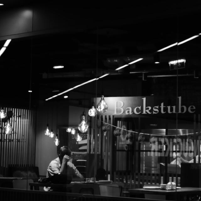 Picture of a man sitting in bakery. He is alone, his right arm is put on the table, the hand at the head like he is holding it up. The left hand is either under the table or on the chair. He might look at a newspaper in front of him. Around him the tables are empty. Above the human is a string of light bulbs, in the background there are shapes of other persons visible.
In the middle of the photo on the left side there are letters, saying "Backstube" - bakery.