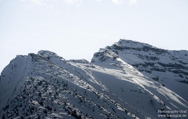 This image captures a breathtaking and rugged snow-covered mountain landscape, evoking a sense of awe, majesty, and untouched natural beauty. The foreground is dominated by steep, jagged peaks blanketed in pristine white snow. The snow's surface is textured with ridges and shadows, highlighting the dramatic contours of the mountains and creating a sense of depth and scale.
At the highest peak, a cross stands prominently against the sky, adding a touch of human presence and spiritual significanc…