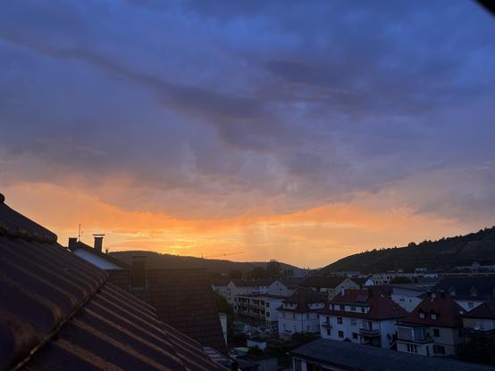 Goldenes Feuer des Sonnenuntergangs über Esslingen; Teile eines Daches und die Stadt sind zu sehen, über dem feurigen Bereich des Sonnenuntergangs am Himmel dunkle Wolken und zu erahnen: Regenfahnen.