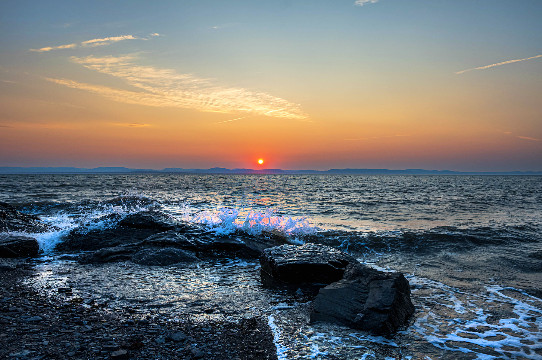 An orange sunset over low hills on the far side of a great river with foamy waves crashing on the rocks in the foreground.
