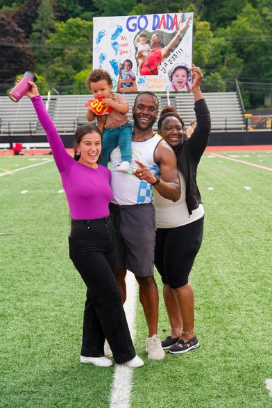 A black man with a baby on his shoulder and a black woman behind him and to the right and a white woman to his front and left hold up a sign that say GO DADDY on astroturf in front of bleachers