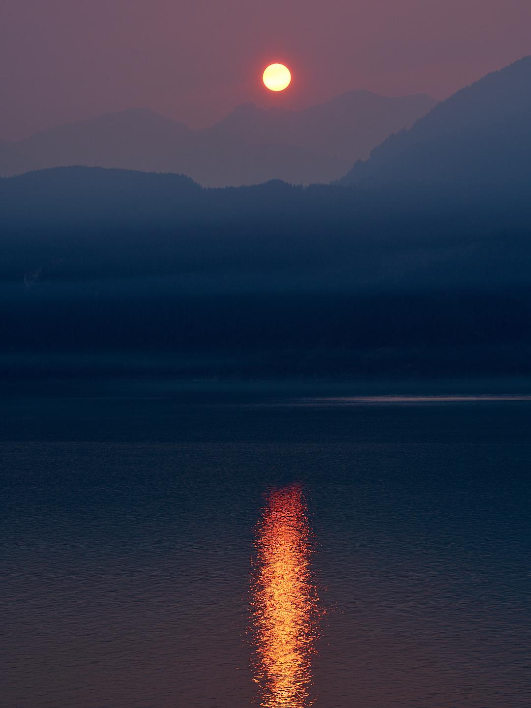 A glowing sun hangs in a smoky purple sky above dark mountain silhouettes, casting a vibrant column of golden light across calm, deep blue water of Pacific Ocean.