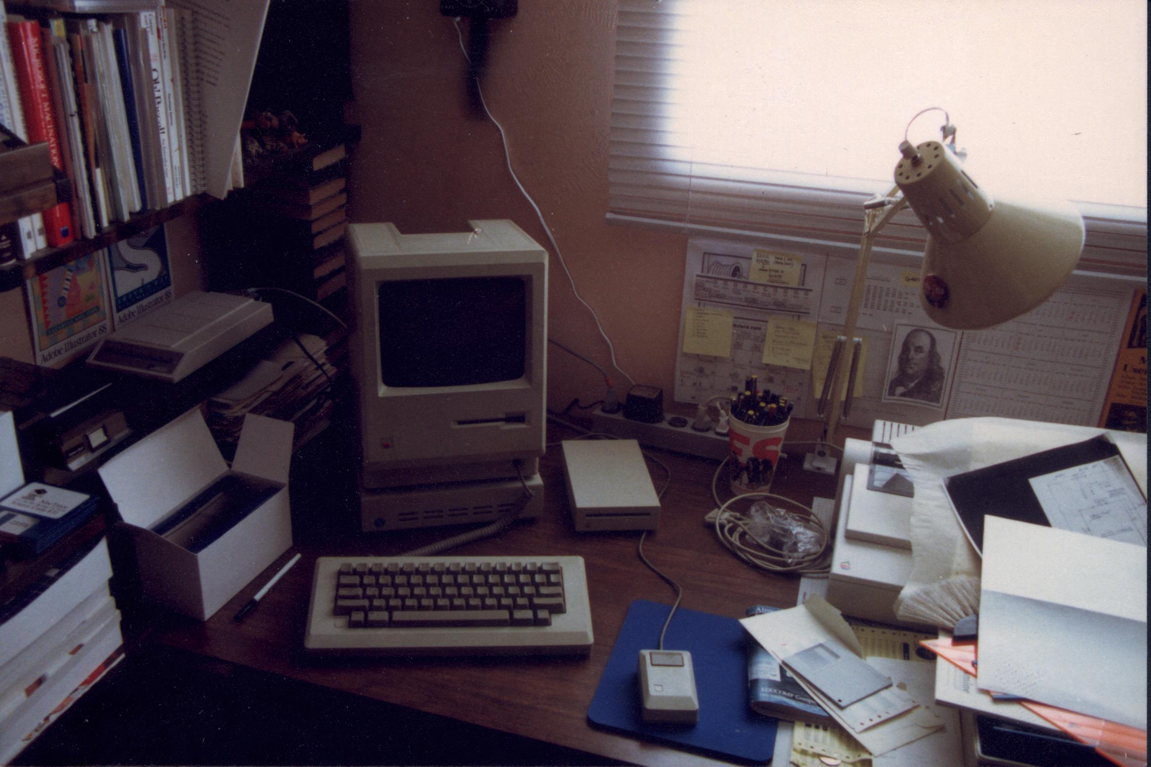A desk with a Macintosh 512K computer, keyboard, and mouse. On the right side of the desk is a pile of clutter that partially obscures the ImageWriter II printer underneath it.