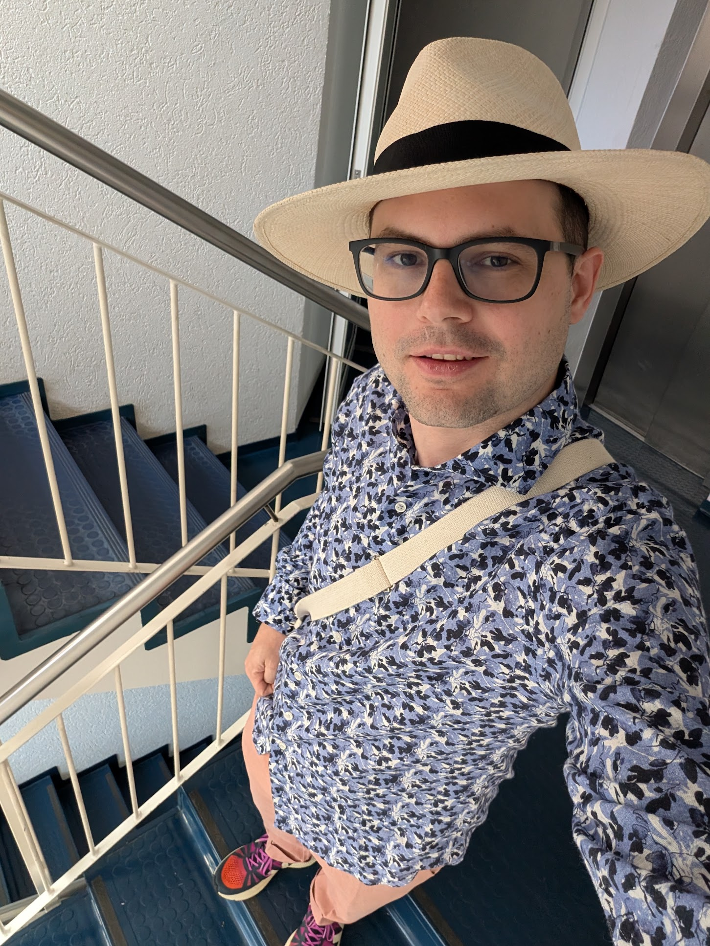 Natural panama traveler hat, blue shirt with leaves, bright rose trousers, and sneakers with a blue/purple/orange kind of transition.