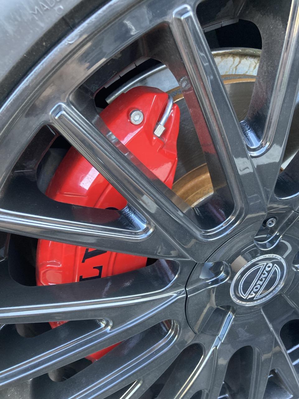 The front left tire of a Volvo.  Inside the spoked hubcap is a medium red, organically rounded brake assembly with a silver band at one end, looking like a mouth.  Above the band is a silver bolt, looking like an eye.  The assembly housing narrows at this end, as a head might.  The whole effect is of a large red fish.