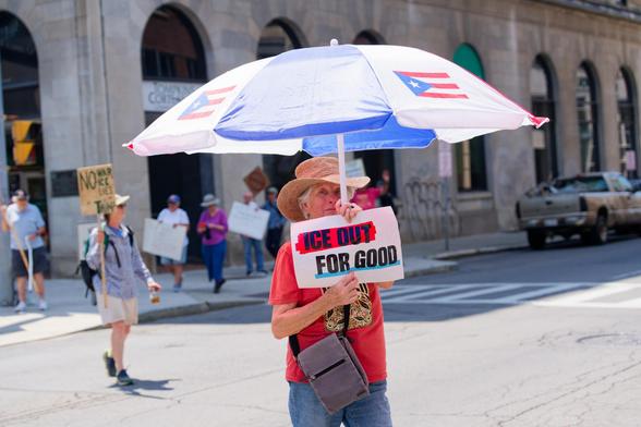 A woman with an umbrella decorated with Puerto Rican flags holds a sign that says ICE OUT FOR GOOD in the middle of a street with more protestors behind