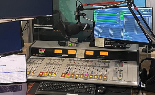 Control board at Vancouver’s Co-op Radio with VU meters, 18 volume faders, several mics headphones and computer screens. 