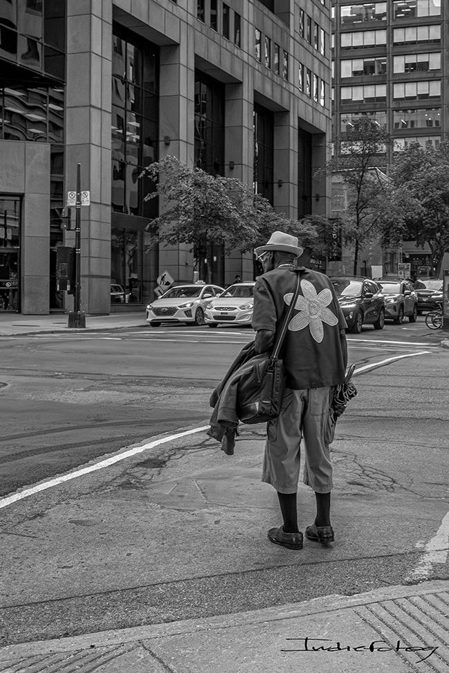 A man is crossing a street wearing wearing 3/4 pants with over the knee socks, a straw hat and a shirt with a big flower on his back.