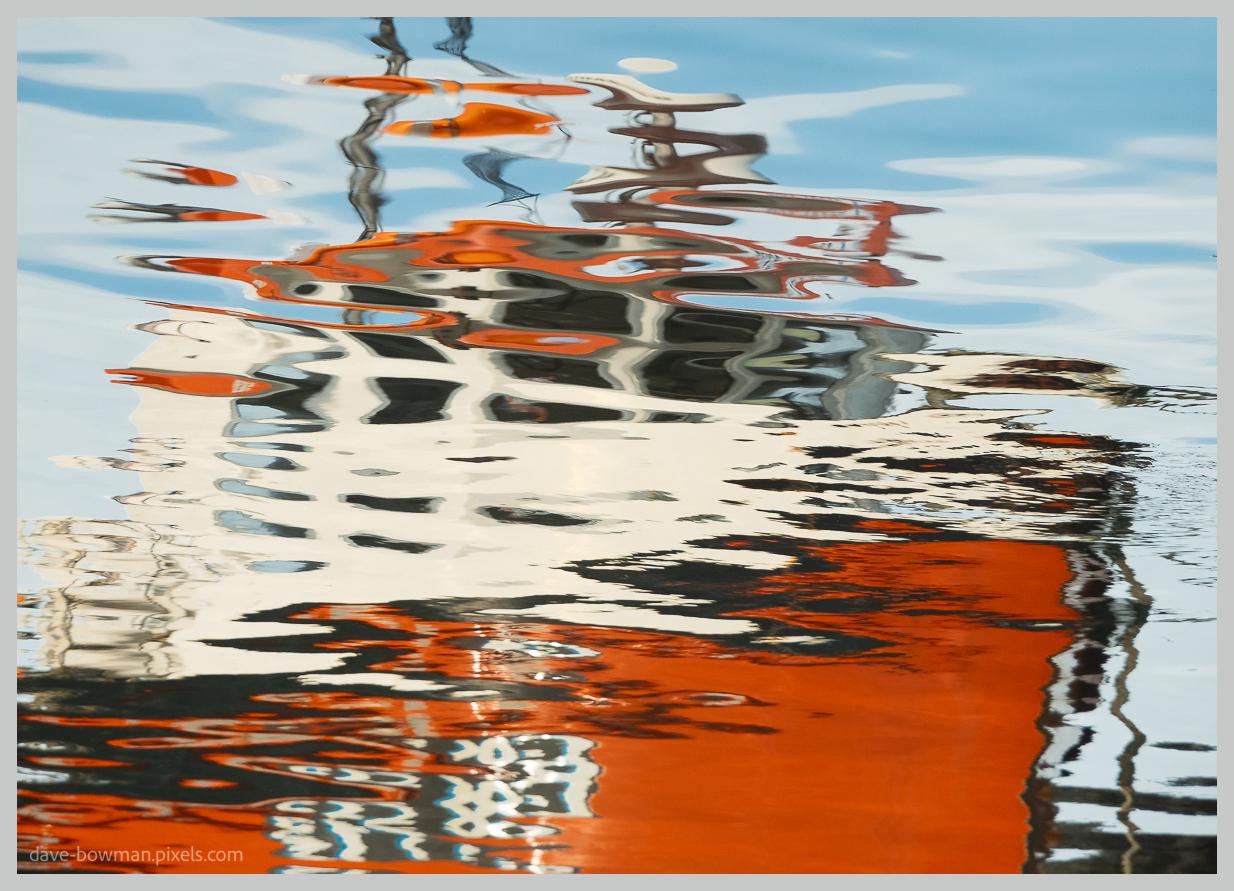 A photograph of a boat reflected in the water at Port Askaig on the Isle of Islay, where the image of the vessel is distorted into broken shapes and flowing lines by the movement of the surface. The reflection appears stretched and fragmented, with colours blending together to create an abstract pattern while still hinting at the boat’s original form. The scene focuses on the changing appearance of the reflection rather than the boat itself, showing how the water transforms a familiar object into something fluid and constantly shifting.