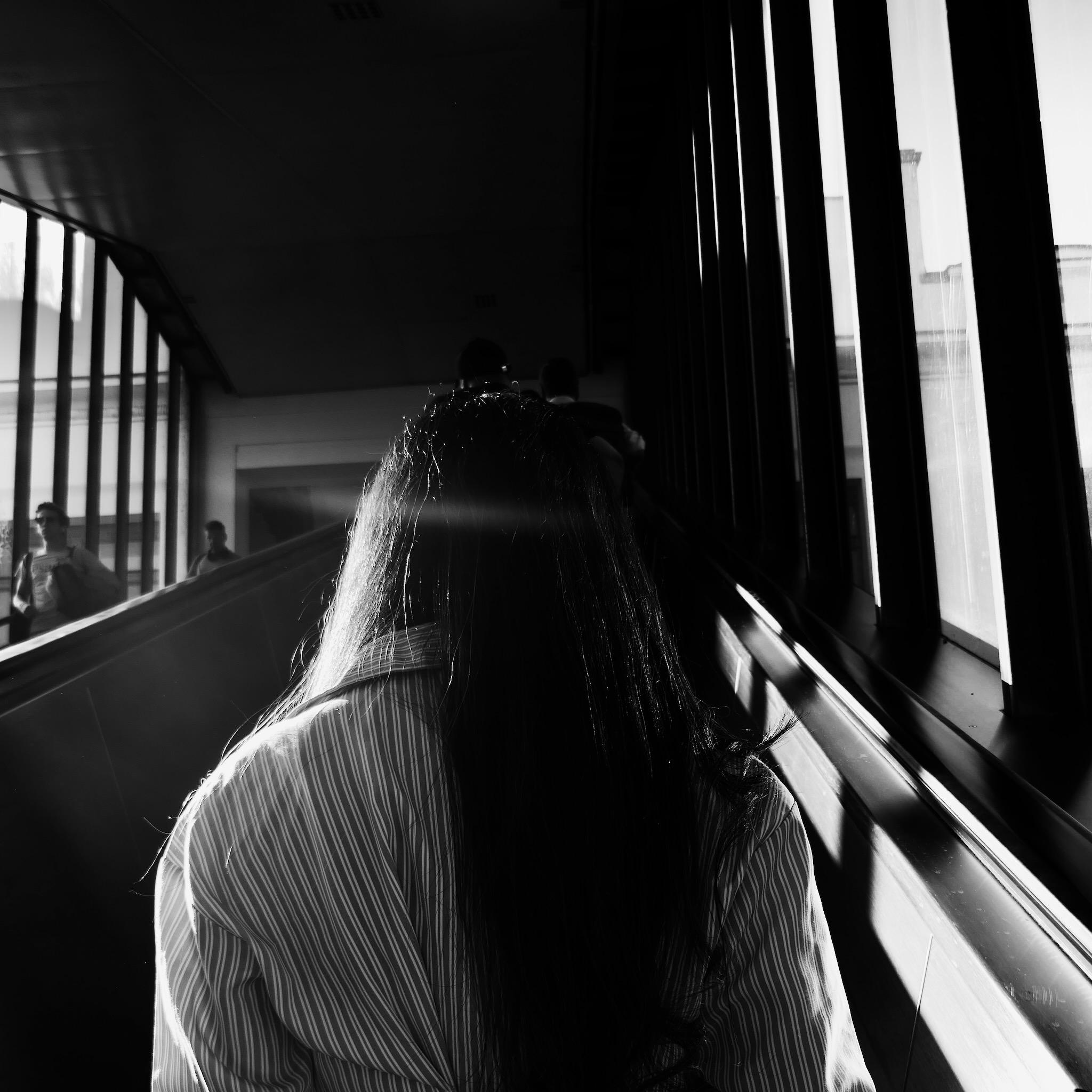 Picture of a young woman on an escalator. She has long hair and a bright dress. Light falls in from the left side and there is a small flare of sunlight breaking on her hair. Otherwise the background is dominated by shadows and dark areas.