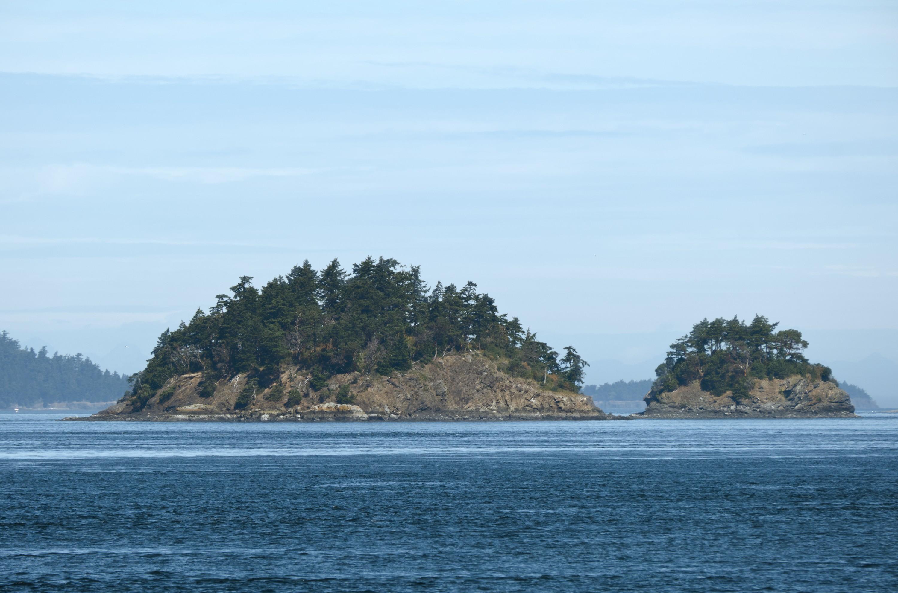 Two small rocky islands at some distance in the middle of a blue seascape with bigger land masses on the horizon. The islands are jutting straight up from the water and capped with forest.