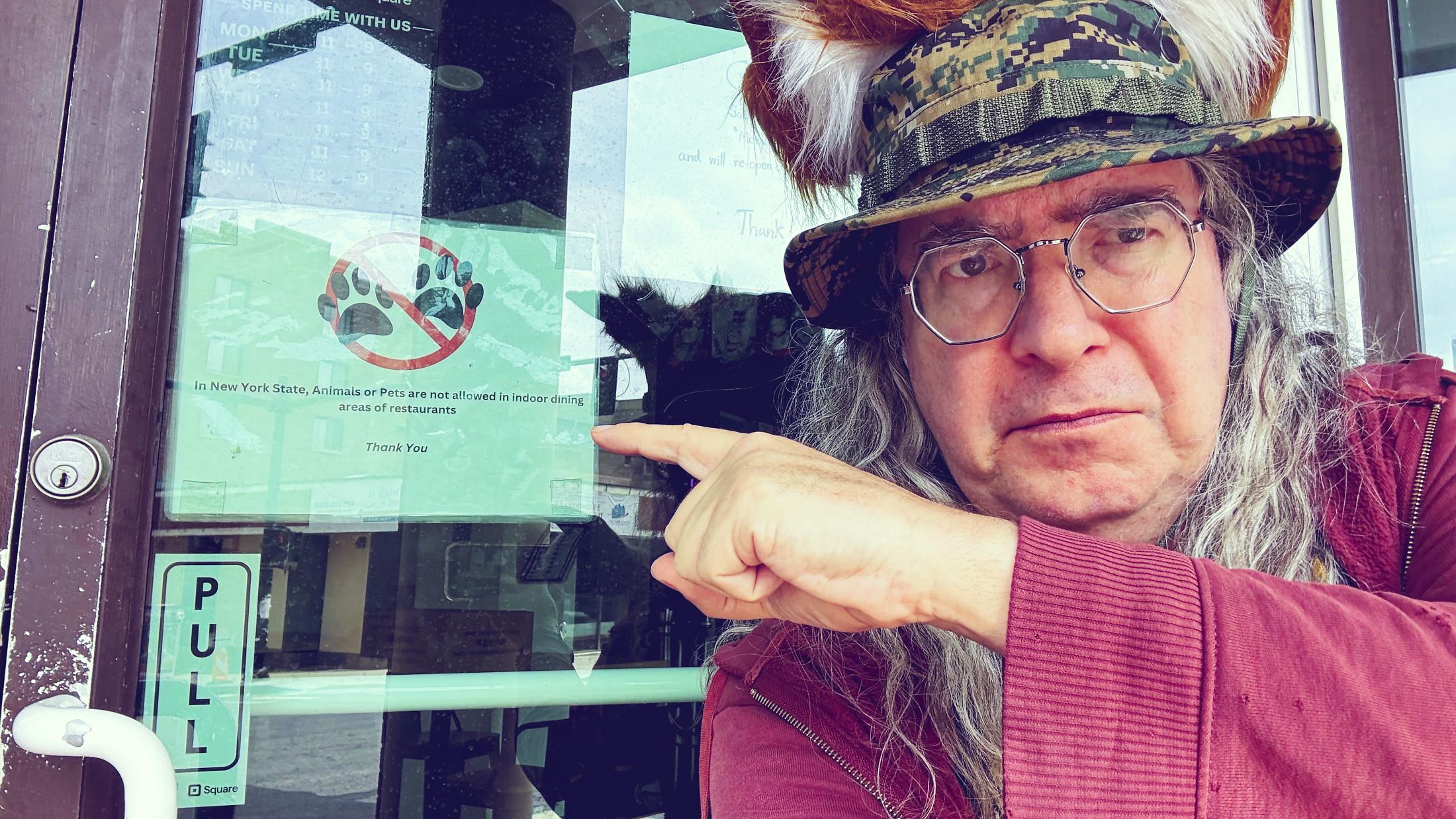 That's me wearing a sour, aged face with a camoflauge boonie cap and fox ears I got off Temu and long curly hair getting dangerously like anime character hair.  I'm wearing an overly well warn maroon hoodie that fits my fall color scheme and i am pointing at a sign at a restaurant with two paws crossed out with a now sign to say that animals are not allowed