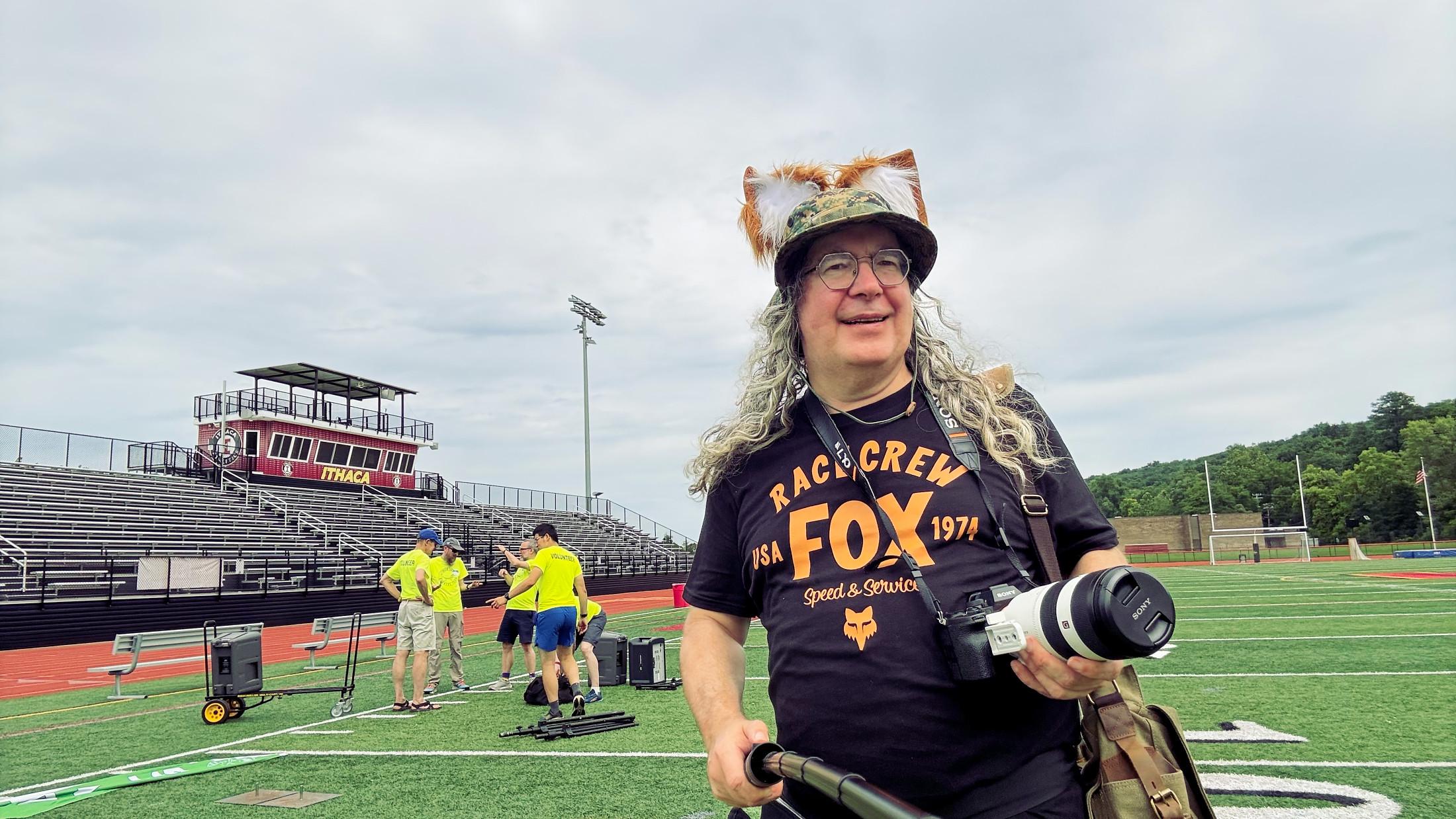 I occupy maybe 15% of the frame with fox ears over a boonie cap with lush long curls cascading past a smiling face with a dark shirt that read FOX RACE CREW and has the Fox Head logo of a legendary motorcross parts company,  I am holding a camera with a big lens and a selfie stick and behind me is a football field with bleachers, a red track,  and yellow-shirted volunteers packing up.