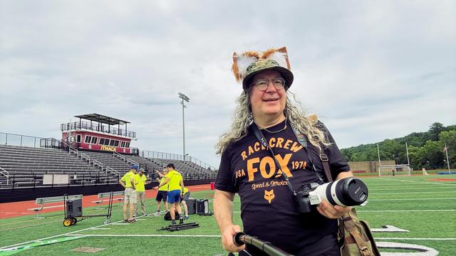 I occupy maybe 15% of the frame with fox ears over a boonie cap with lush long curls cascading past a smiling face with a dark shirt that read FOX RACE CREW and has the Fox Head logo of a legendary motorcross parts company,  I am holding a camera with a big lens and a selfie stick and behind me is a football field with bleachers, a red track,  and yellow-shirted volunteers packing up.