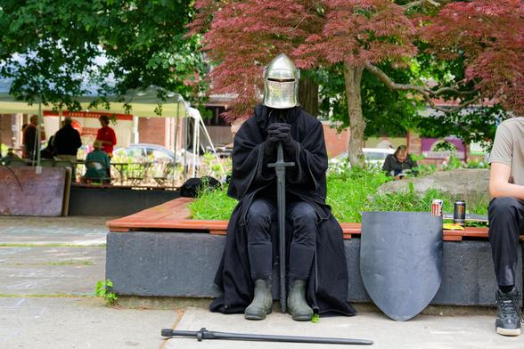 A man with a metal knight mask and black cloak as well as two swords and a shield poses on a bench surrounding a flower bed