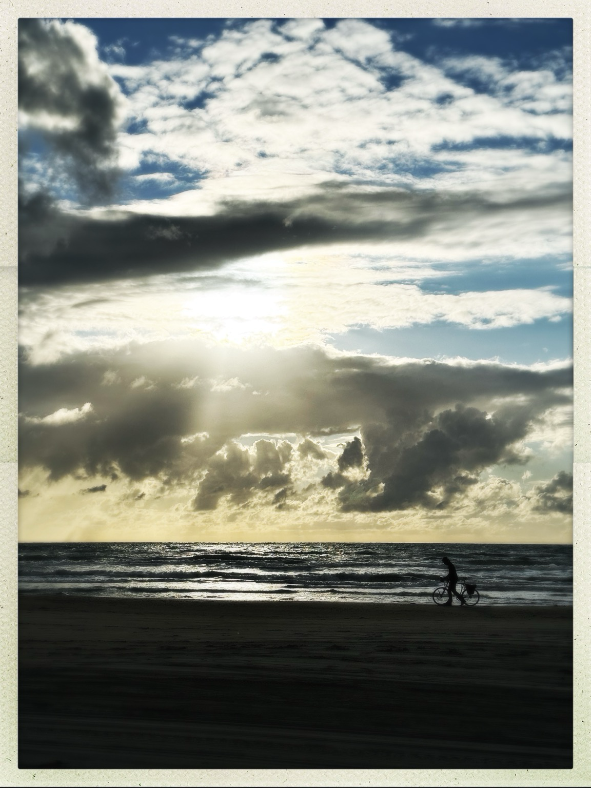 Das Bild zeigt einen Strand in Dänemark. Am Wasser steht ein Radfahrer und genießt den Blick über das Meer und einen freundlich bewölkten Abendhimmel. Auf das Bild ist ein Filter gelegt, es wirkt ein wenig wie ein altes Polaroid mit einem weißen Rahmen.