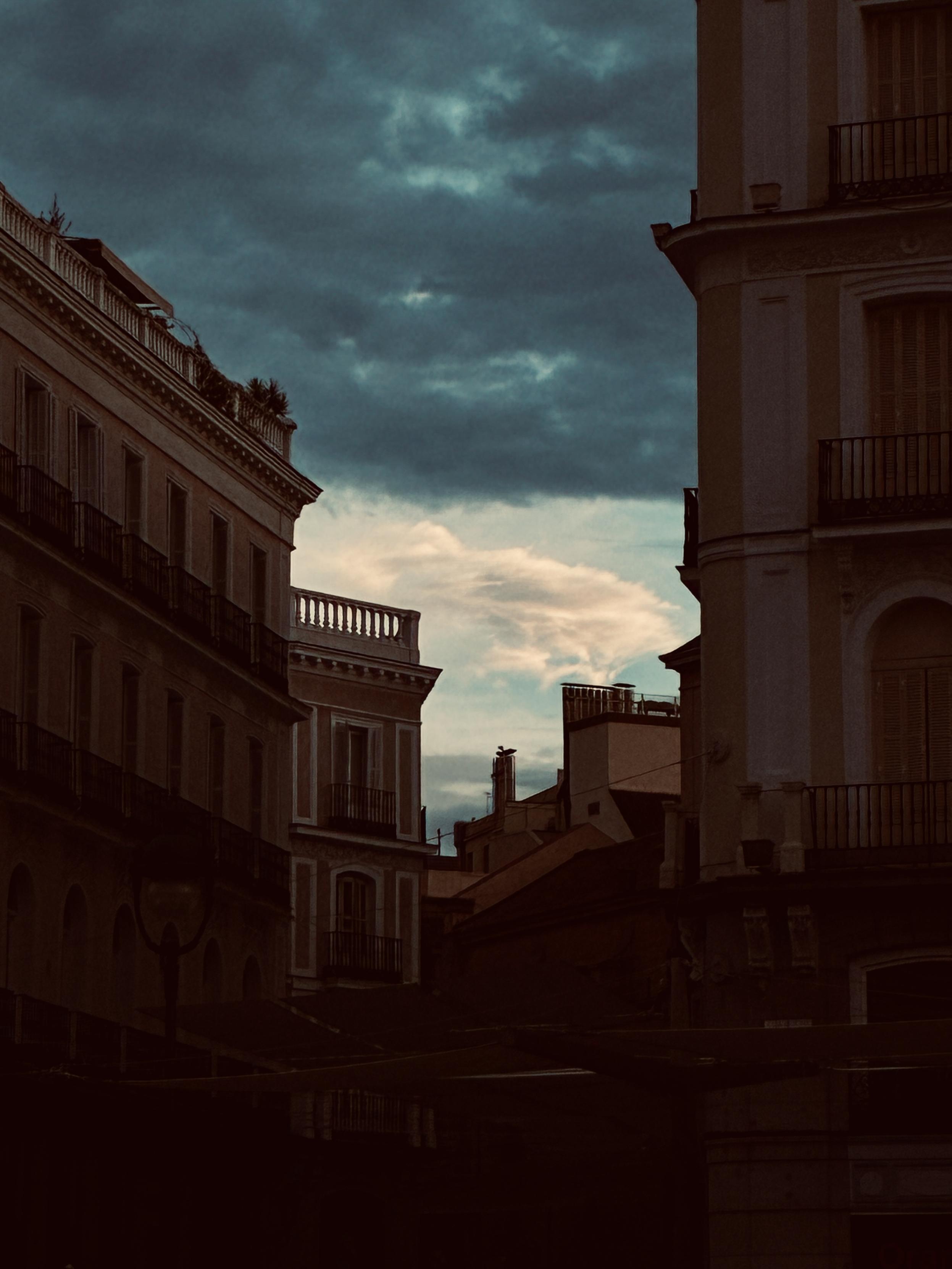 Fotografía de un cielo parcialmente nublado al amanecer, entre edificios dispuestos aleatoriamente. Algunas nubes son blancas, con tonos anaranjados, y otras azuladas y oscuras. Los edificios, todos rojos.
