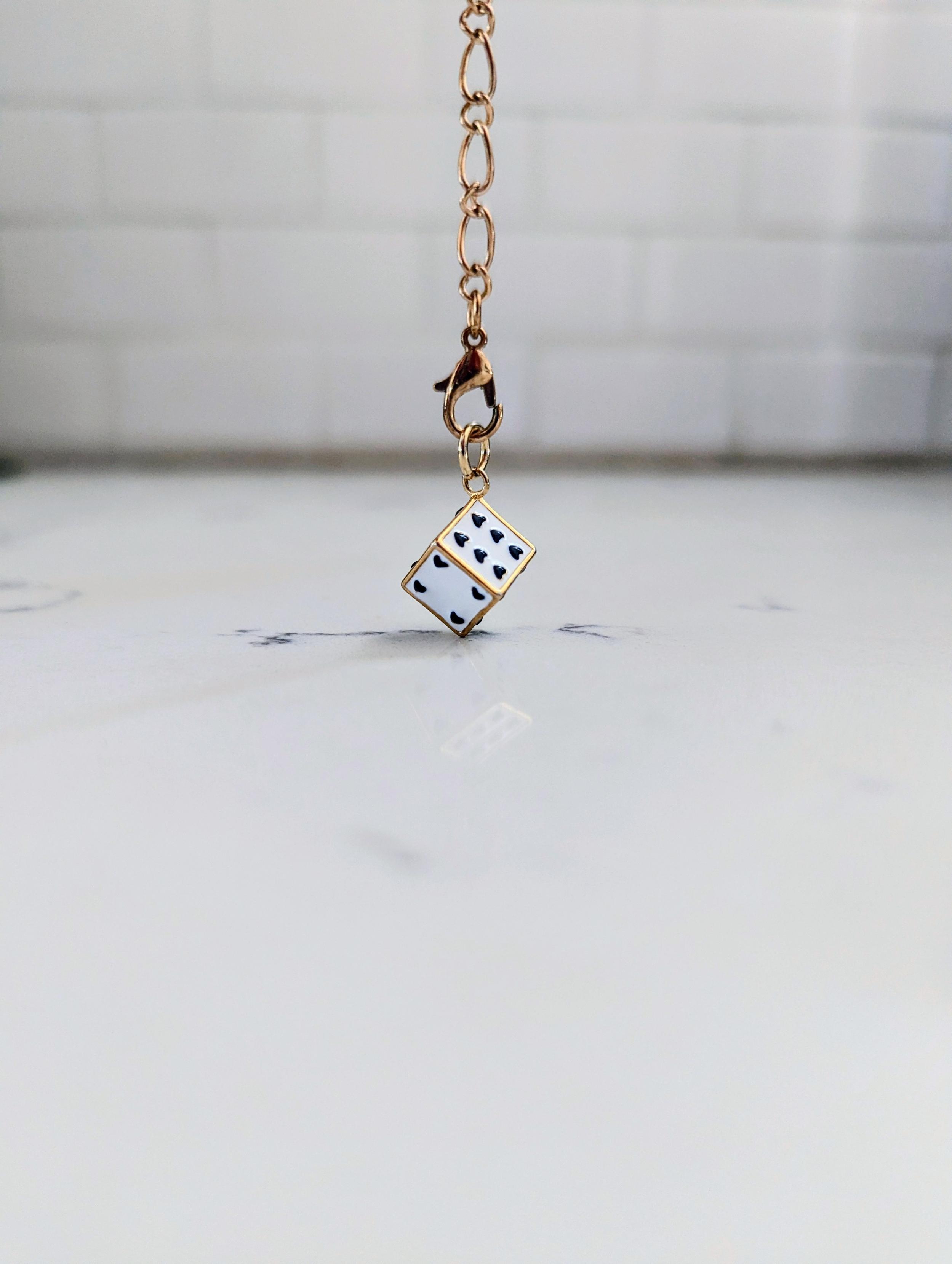A photo of a small enamel charm that's a white die where the dots are hearts, dangling from a gold chain