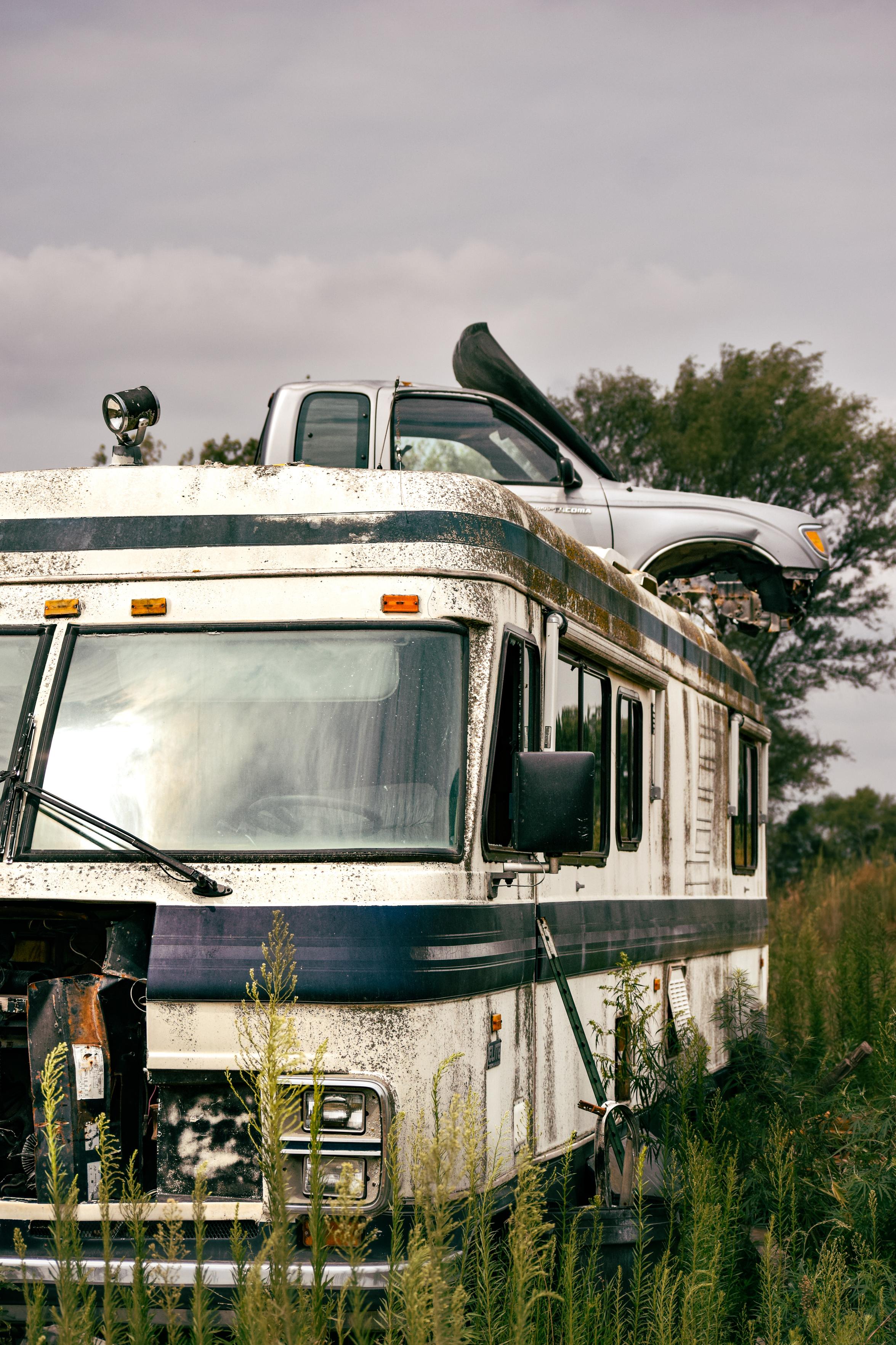 Weathered RV in overgrown field, cloudy sky, pickup truck on roof. Scene feels abandoned, evokes nature reclaiming man-made objects.