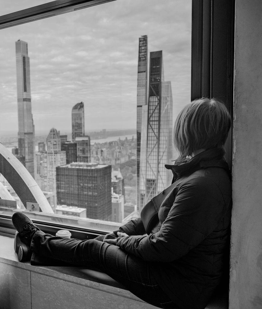A black-and-white photo of a person sitting in a large window well, seen from the side, resting with one leg up and a coffee cup beside them. They look out through the glass at a city skyline of tall skyscrapers under an overcast sky.