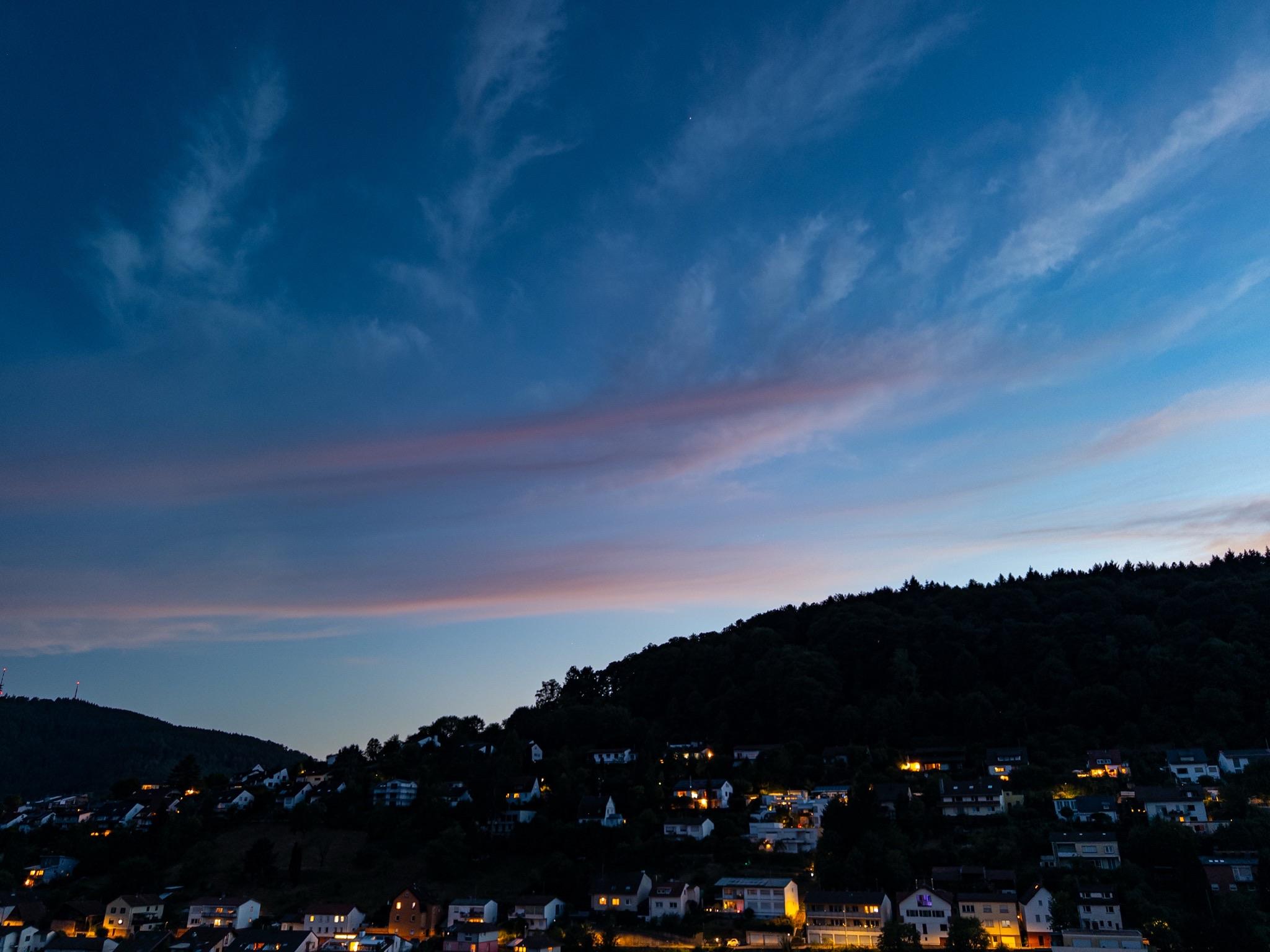 A view across a valley about forty minutes after sunset. The hills are dark with white houses showing orange lights. The sky is blue with some horizontal pink-grey clouds and tails of white above.