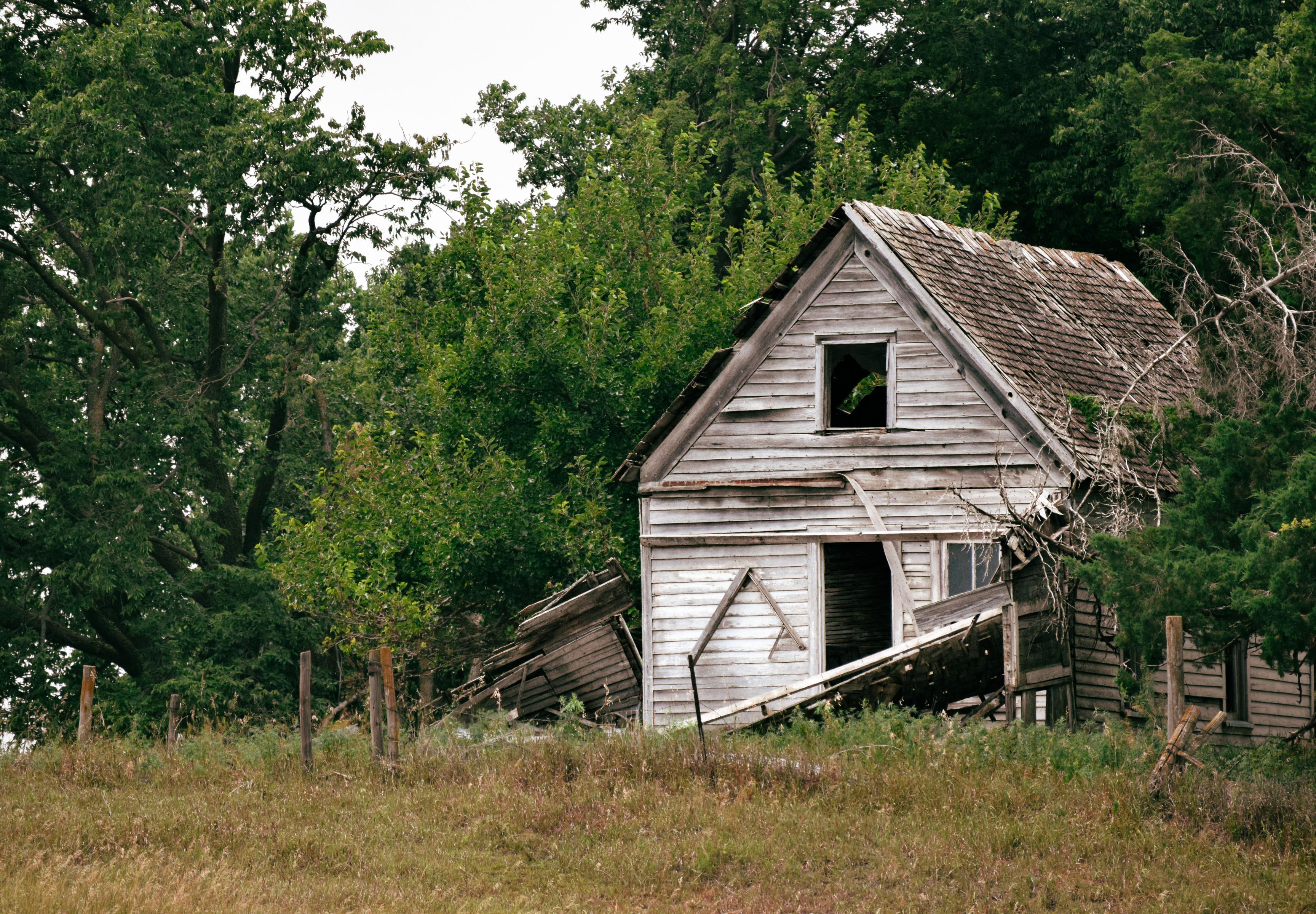 An old, abandoned wooden house with a collapsed porch sits in a grassy field, surrounded by dense, green trees, conveying a sense of decay and solitude.