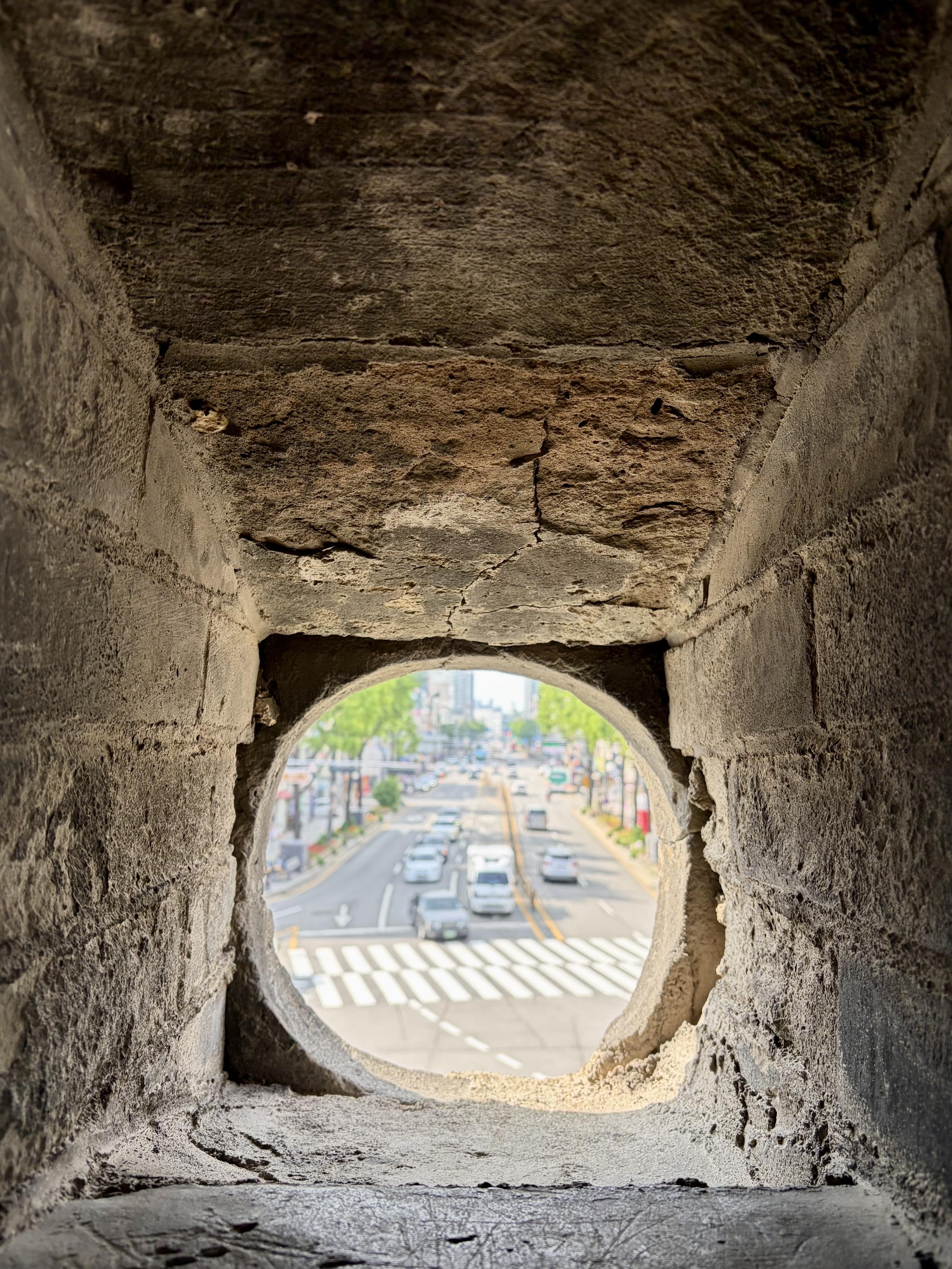 The view from a hole in a fortress wall looking down at a city street. Cars are driving  on the street that is lined with trees and buildings.