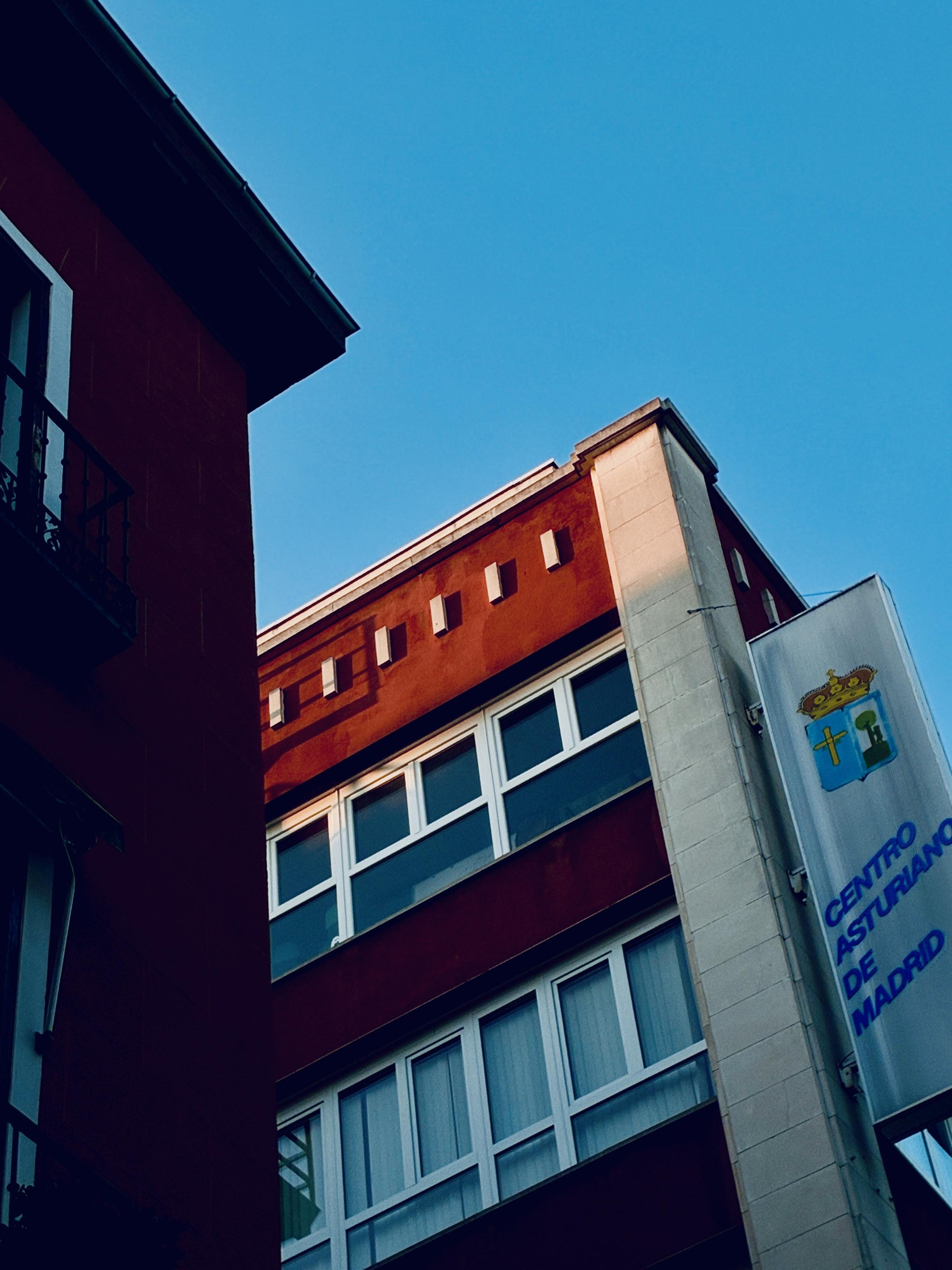 La parte superior de dos edificios crean una figura geométrica entre triangulo o rectángulo con un lado diagonal. El sol ilumina la azote de uno de ellos, en el que reza, en una pancarta, “Centro Asturiano de Madrid”.