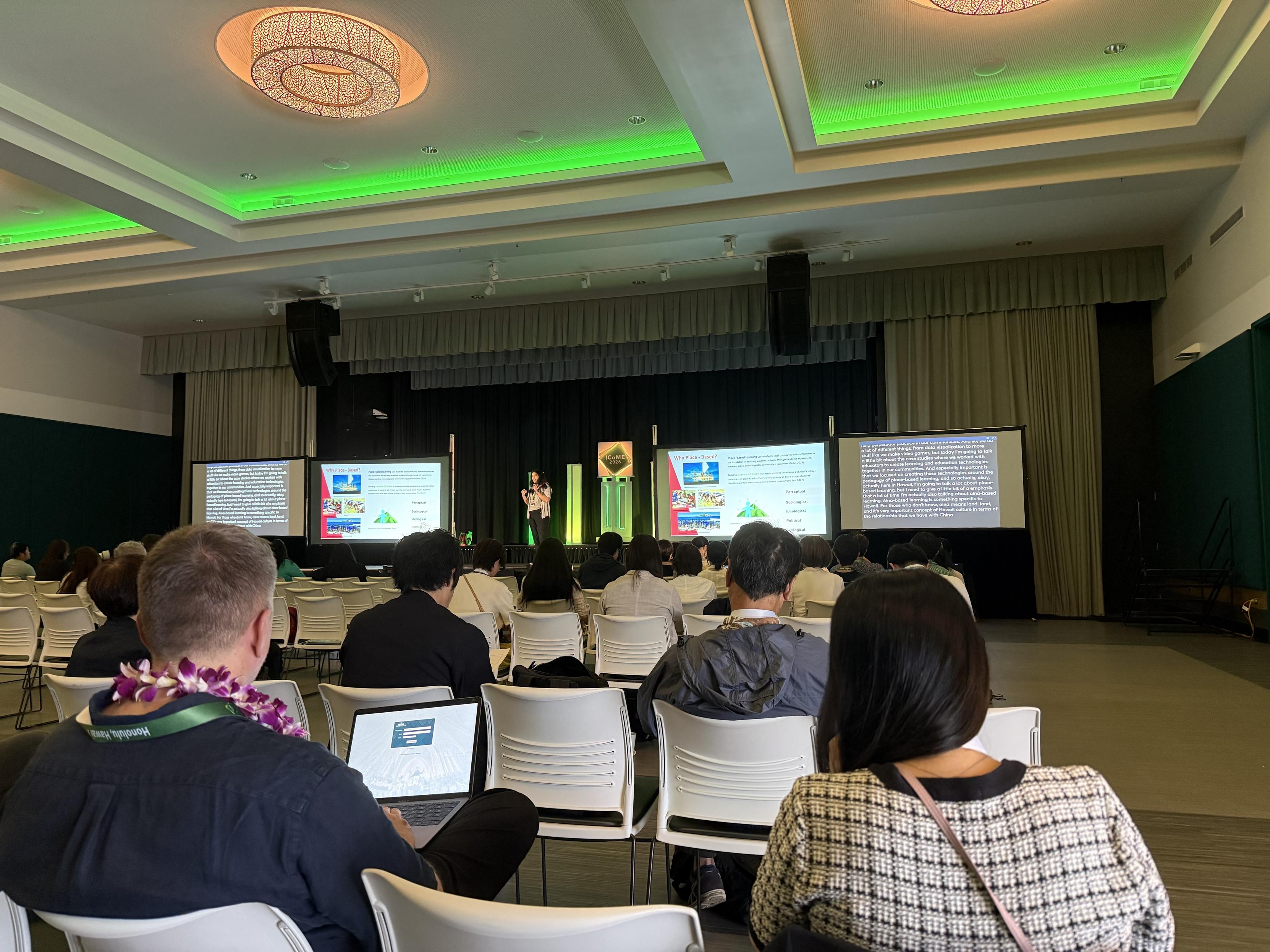 A presentation is taking place in a conference room filled with attendees. A speaker stands on stage in front of two large screens displaying text and images related to the topic. The audience is seated, some with laptops, and greenery accents are visible in the room.