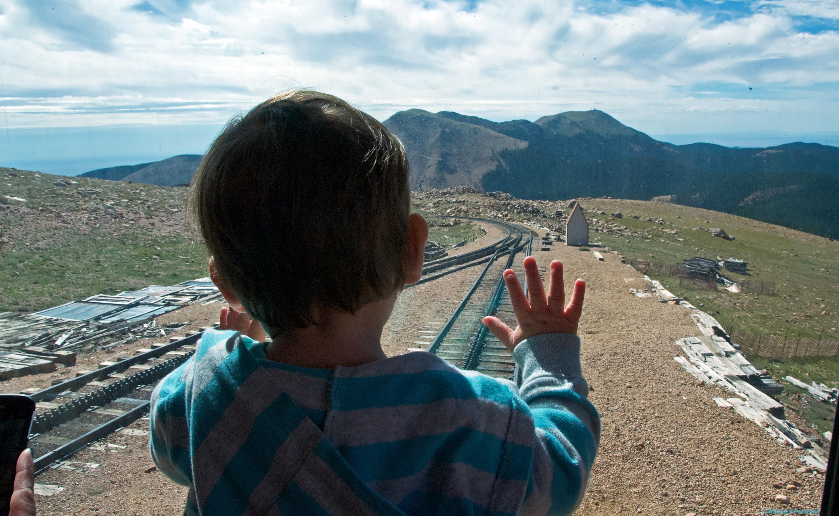 Photo by Fenichel

A young boy has his hands on the windshield in front of him, at the front of a cog rail train heading down the Rocky Mountains from Pike's Peak. Most of the center is the boy from behind, in a fuzzy blue sweater, mostly in silhouette, as there is a bright day in front of him, with the cog tracks ahead, grass (and marmots) off to the right, and mountains straight ahead, headed down..