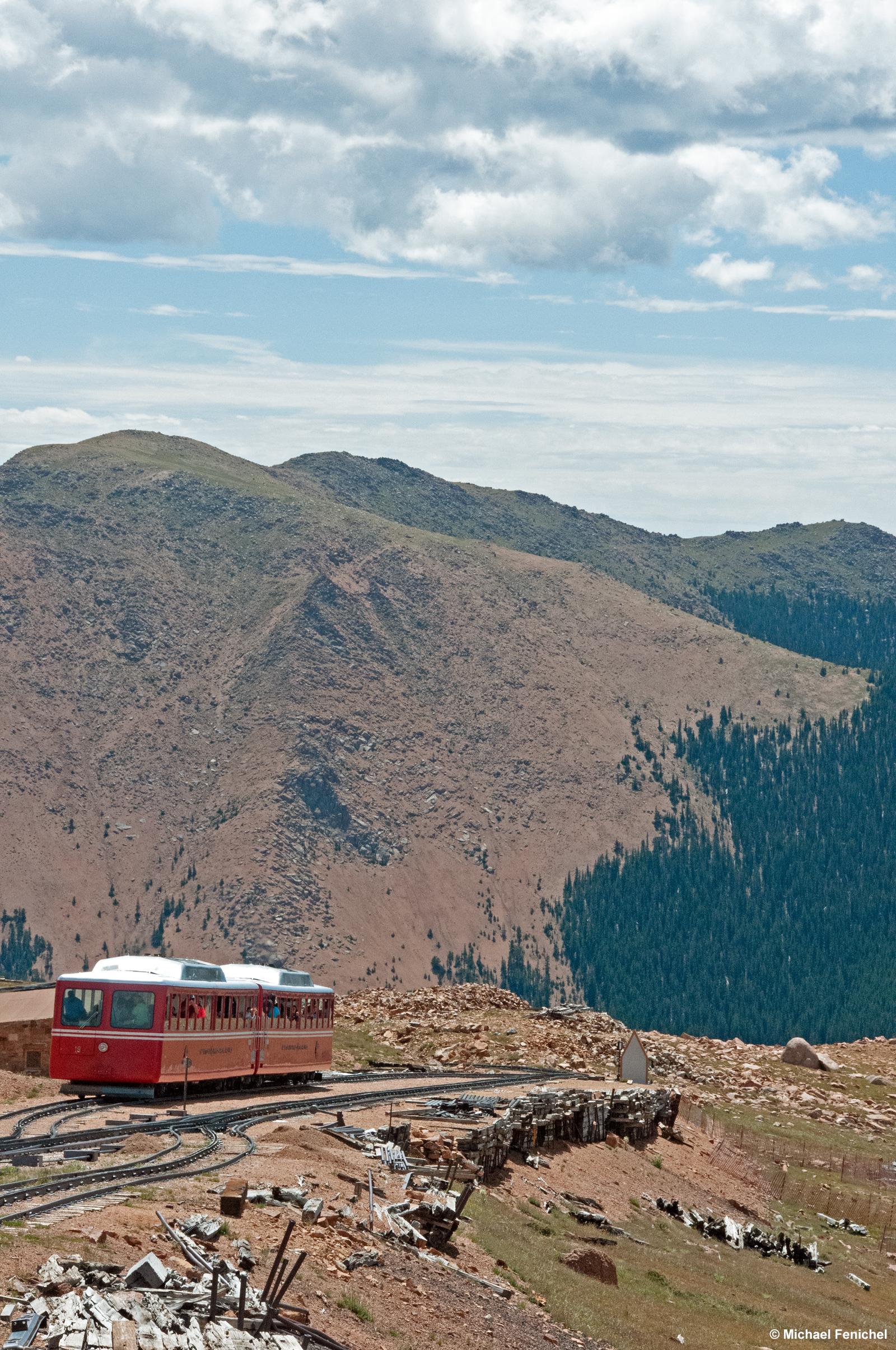 Photo by Fenichel - 
View of a red cog train heading up the Rocky Mountains (Front) towards Pike's Peak. Behind the approaching train are mountains, and before it and behind, are the cog tracks, including the track-switch that this one is waiting at, as our cog train heads downward, at which point the other train will switch to the track to the top.