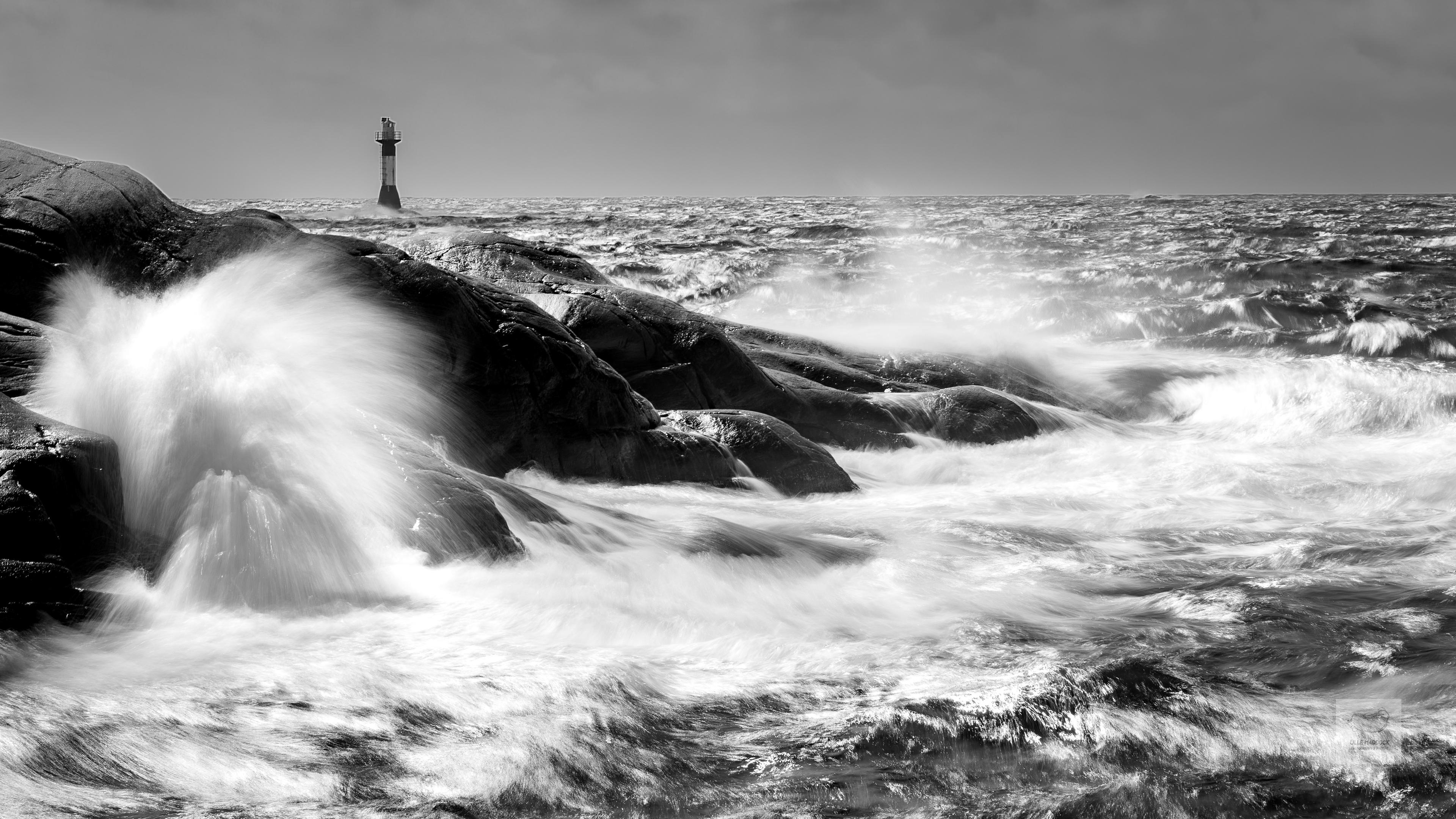 Waves crashing over black rocks with a lighthouse in the background