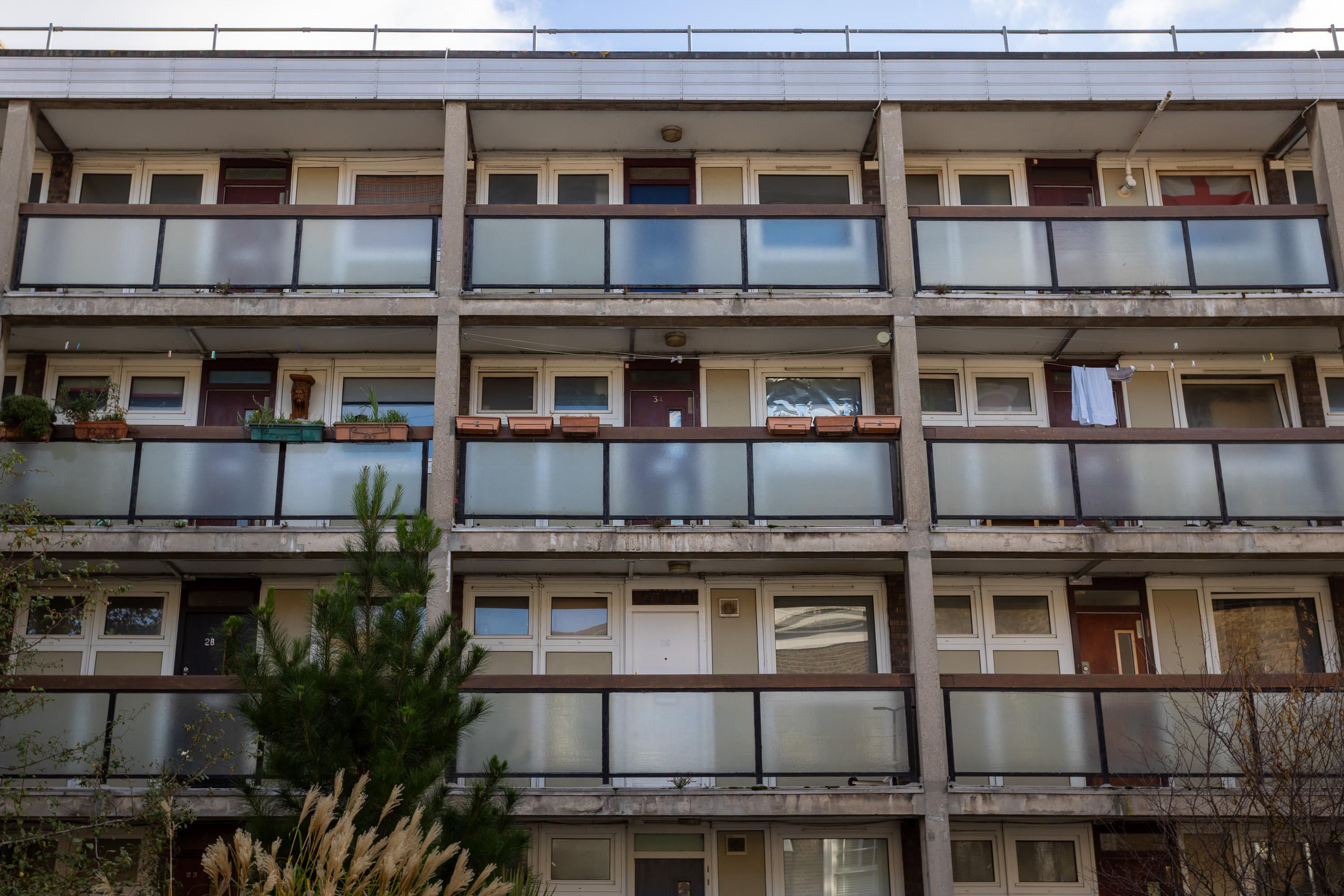 A three-storey block of flats with frosted glass balcony railings, white doors, and windows. Some balconies have planters, potted plants, or washing hanging. There is greenery, including a pine tree, in front of the building. The sky is partly cloudy.