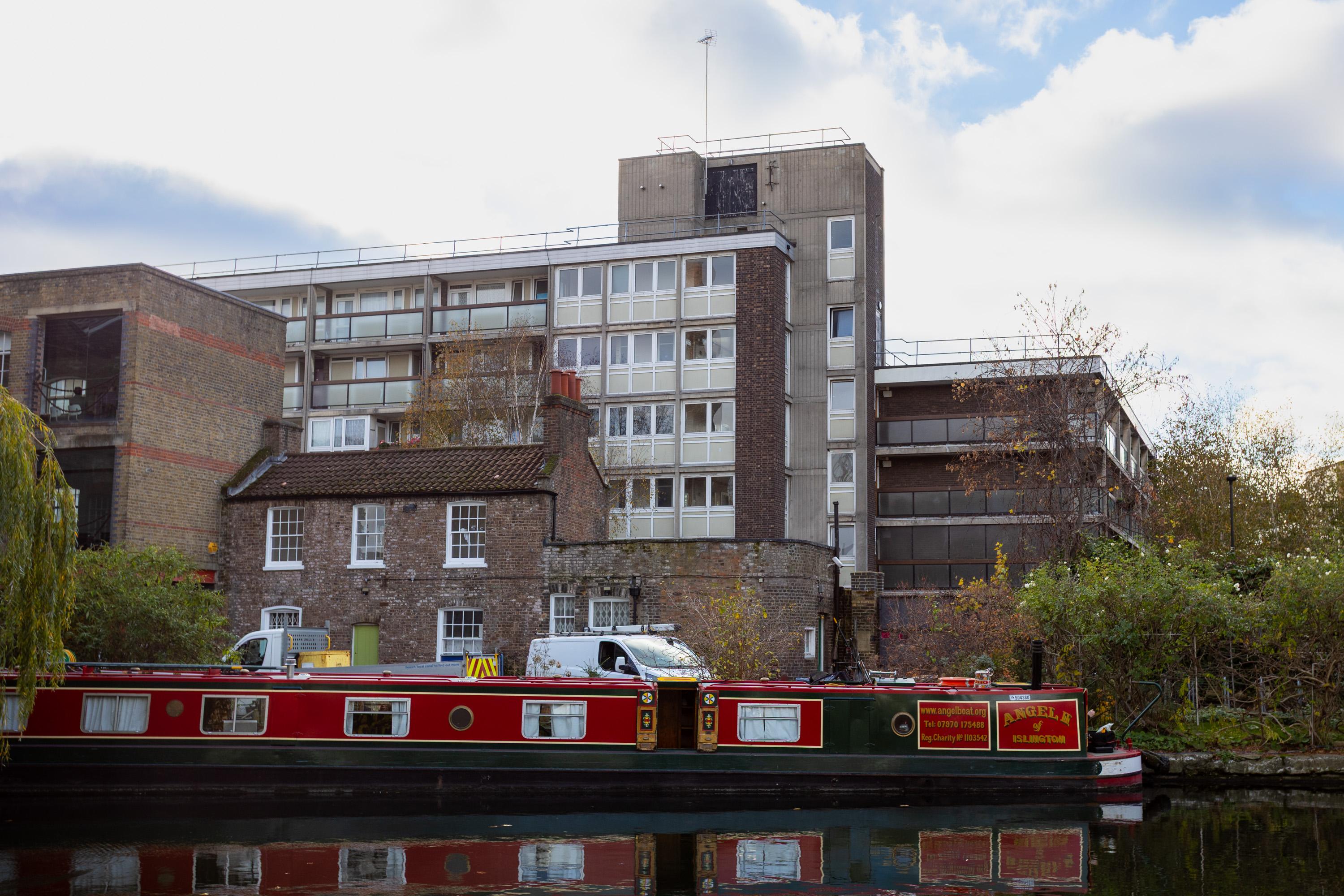 A red canal boat floats on a calm canal in front of a two-storey brick cottage and a tall, modern block of flats. Trees with autumn leaves border the water, and a white van is parked beside the cottage. The sky is cloudy.