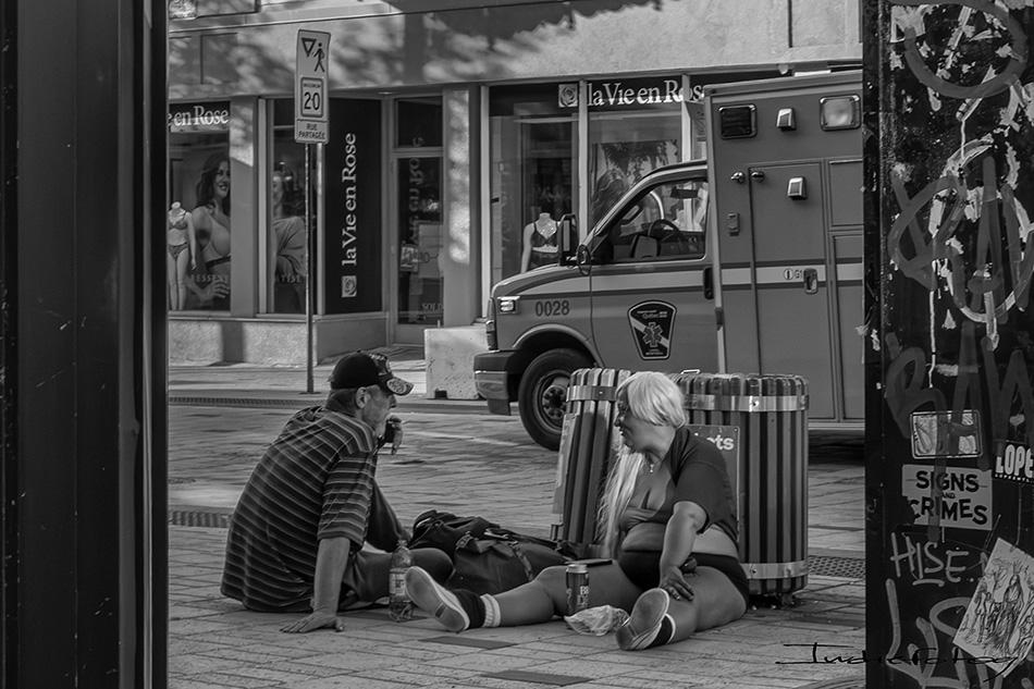 Two street characters sitting on the sidewalk, beside a trash can , with a big store sign i the back saying: "La Vie En Rose"