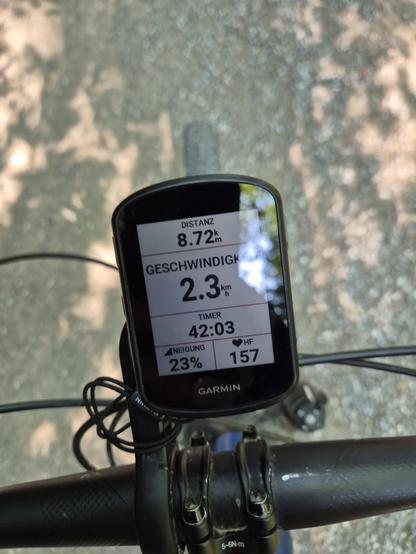 The photo shows a close-up of a Garmin cycling computer mounted on a bicycle handlebar. The screen displays the following metrics in German:
- Distance (Distanz): 8.72 km
- Speed (Geschwindigkeit): 2.3 km/h
- Timer: 42:03
- Incline (Neigung): 23%
- Heart Rate (HF): 157 bpm
The background is blurred, showing a sunlit path with tree shadows, suggesting the ride is taking place on a forest trail or park. The device is securely attached, and cables are visible, likely for sensors or charging. The i…