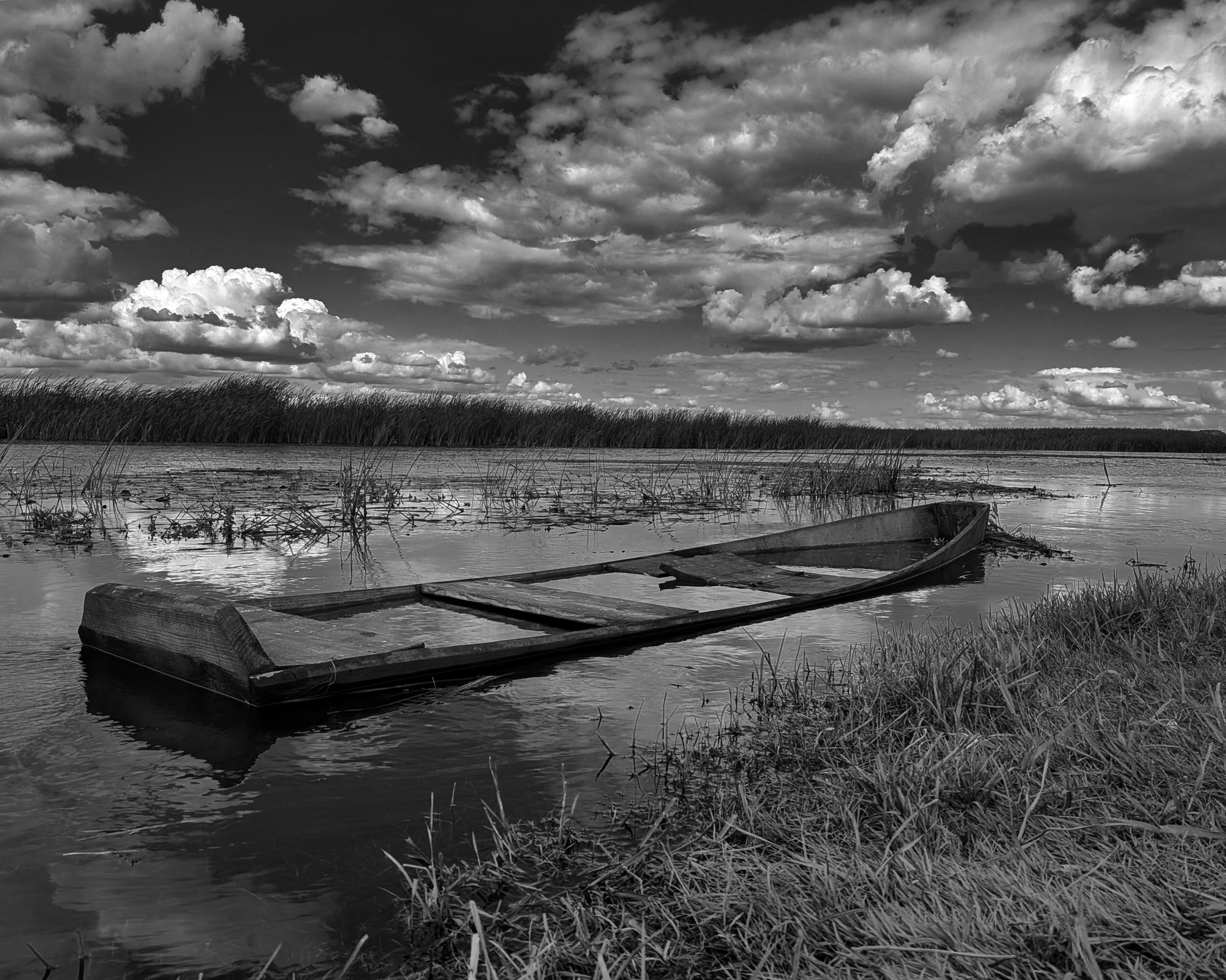 Black and white photograph of an old wooden rowboat resting in shallow water at a river's edge, surrounded by reeds, with a dramatic cloudy sky above.
