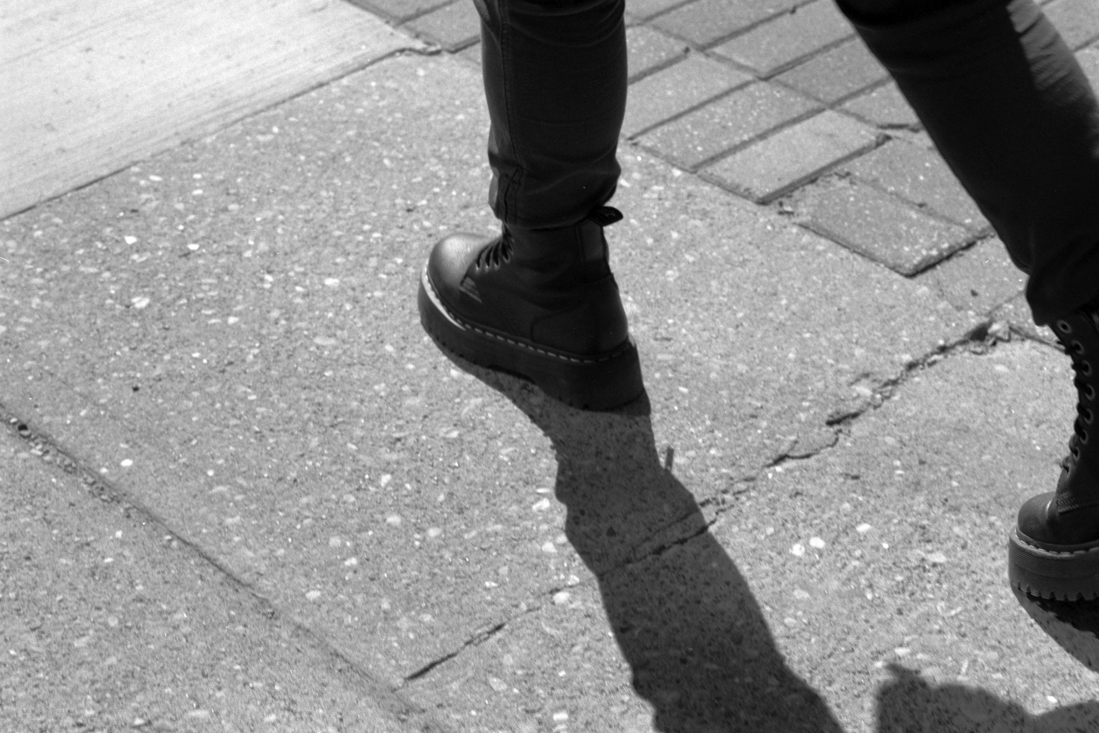 A black and white 35mm photograph of a woman walking, mid-stride.  She is wearing black boots and black jeans and her leg is casting a shadow on the pavement.
